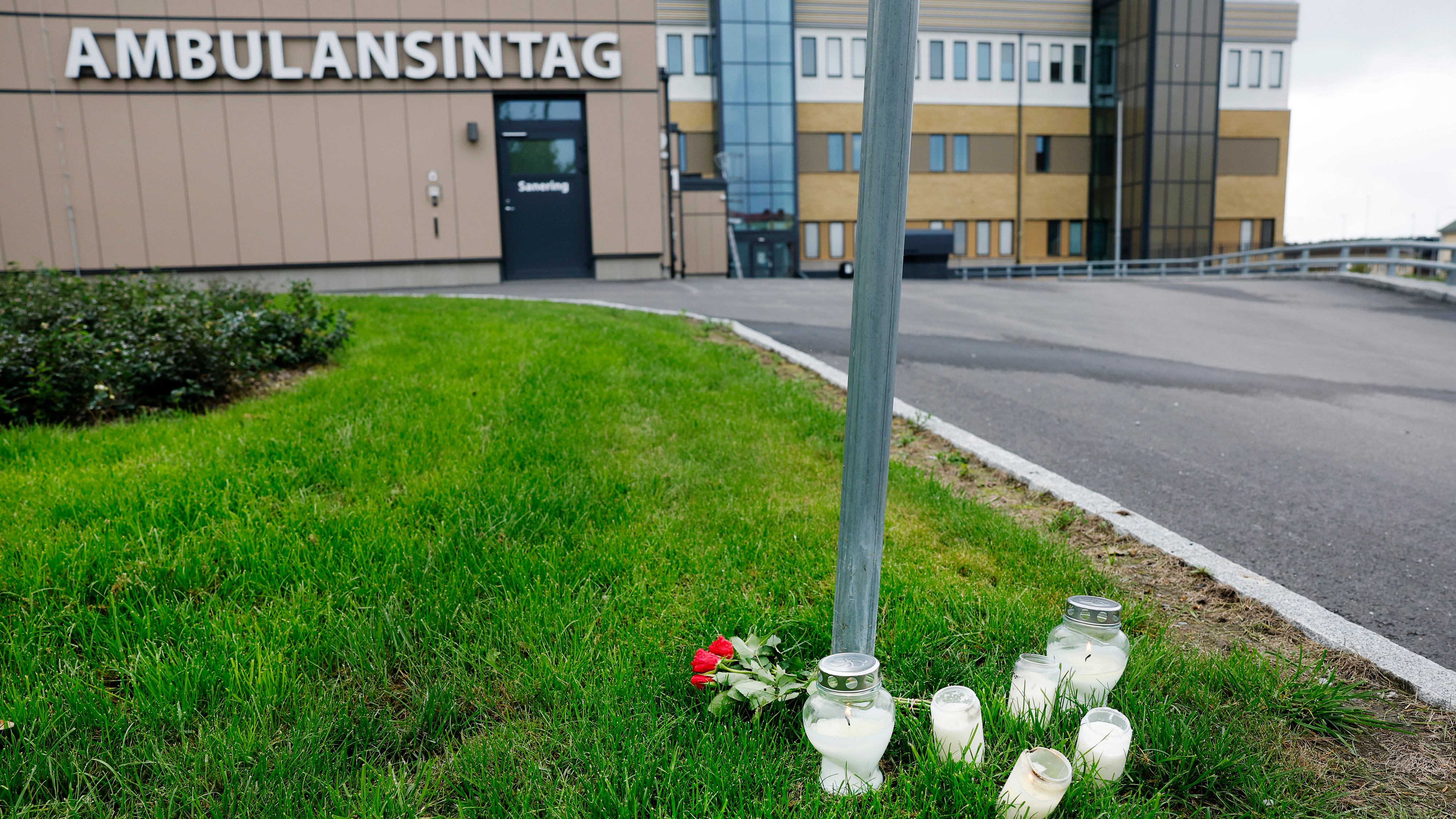 Flera ljus och röda rosor står på gräs nära en skylt vid en byggnad med skylten "AMBULANSINTAG".