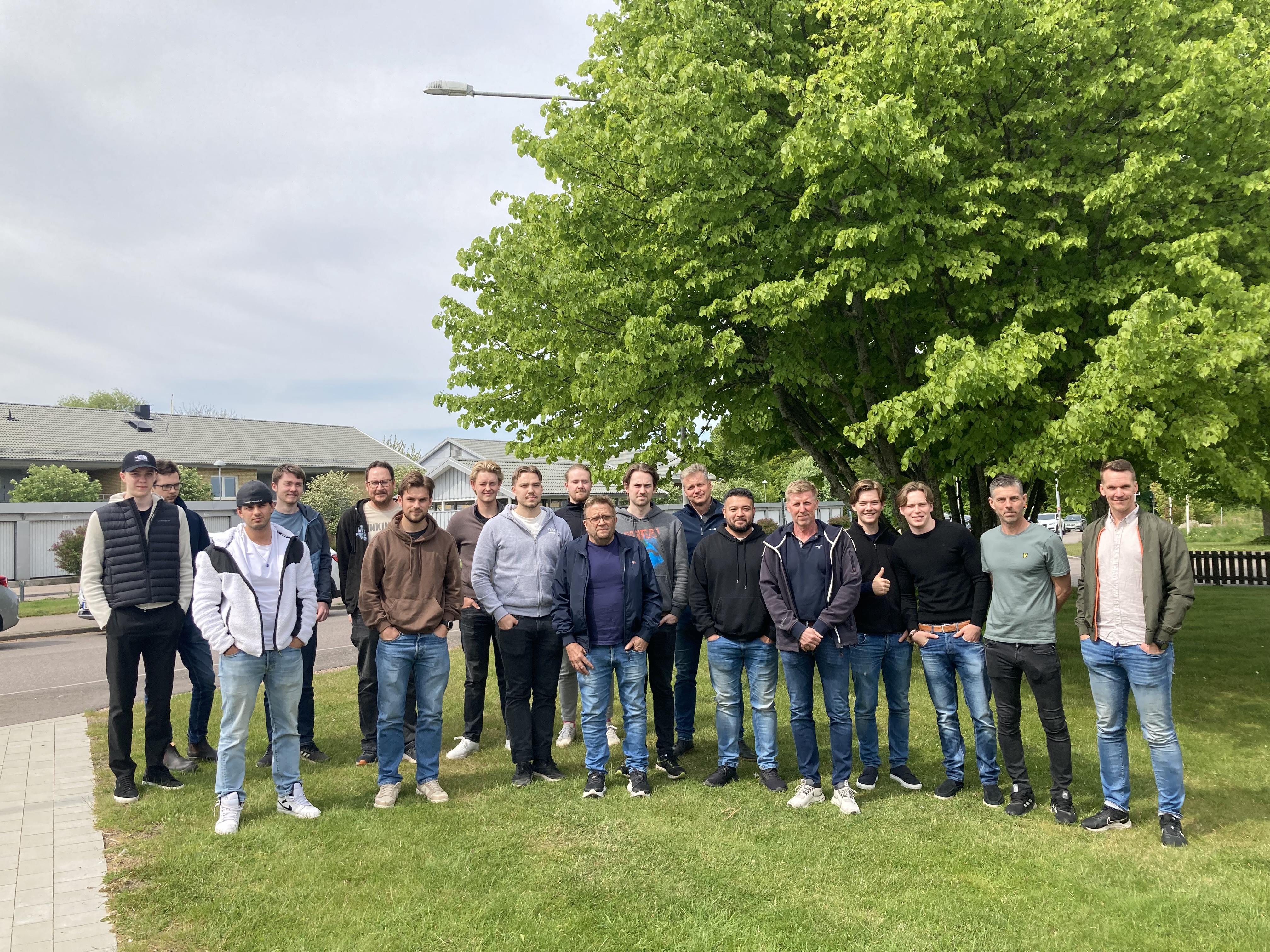 Sexton personer står på en gräsmatta framför ett träd, med hus och himmel i bakgrunden.