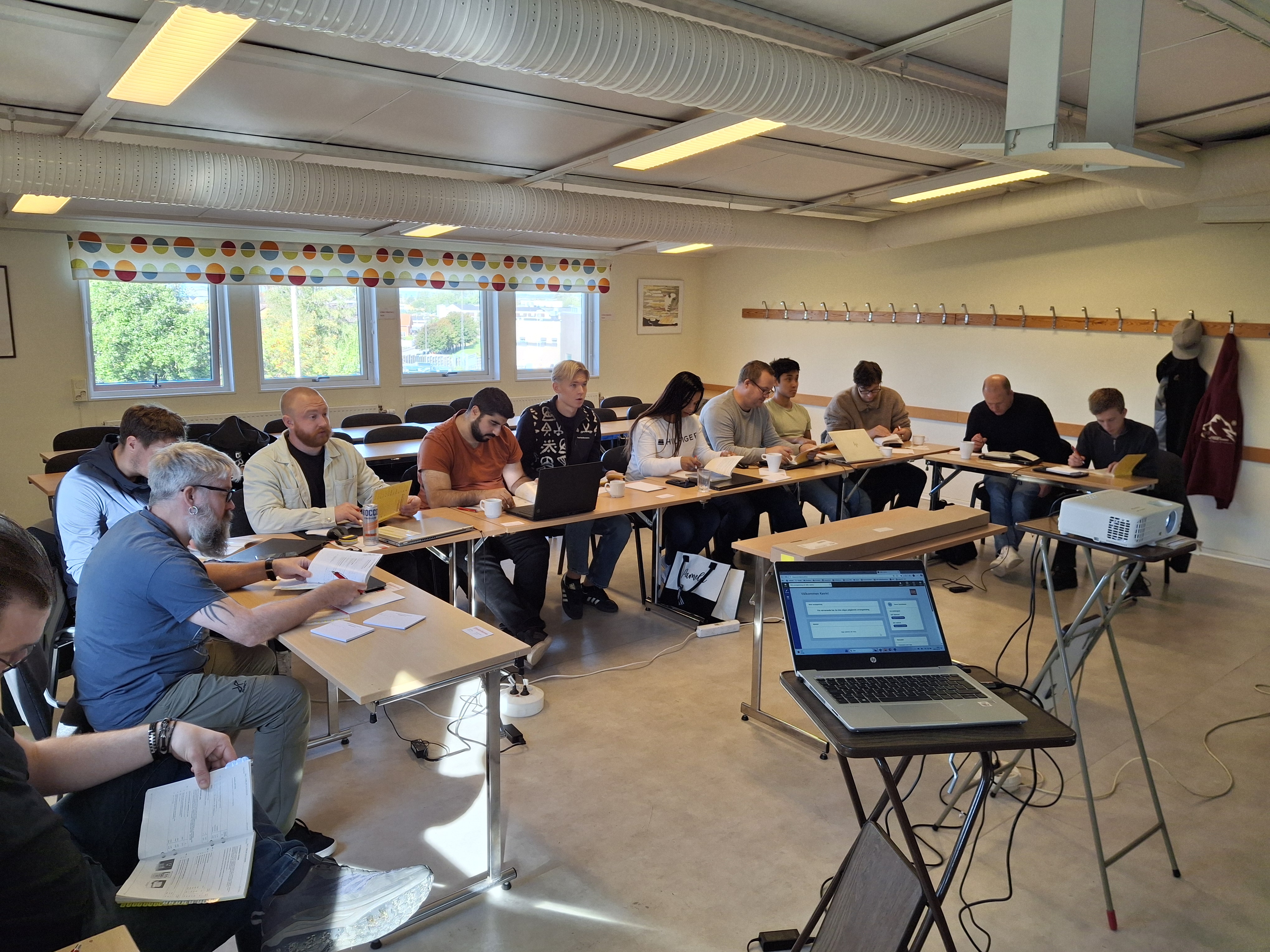 Personer sitter runt bord i ett klassrum, arbetar med papper och använder bärbara datorer, en projektor visar information framme.