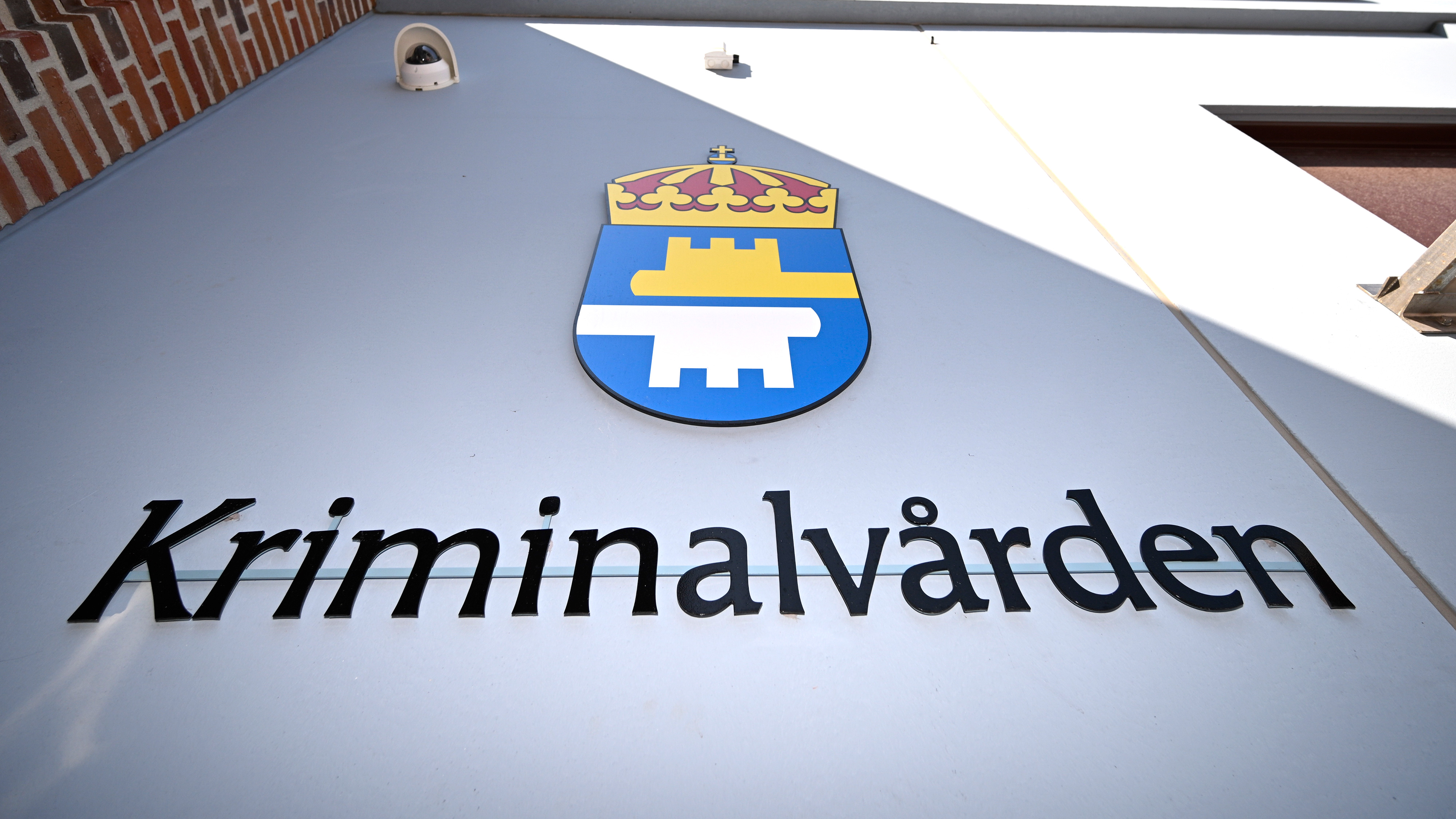 En vägg med Kriminalvårdens logotyp och namn i svarta bokstäver under en säkerhetskamera.