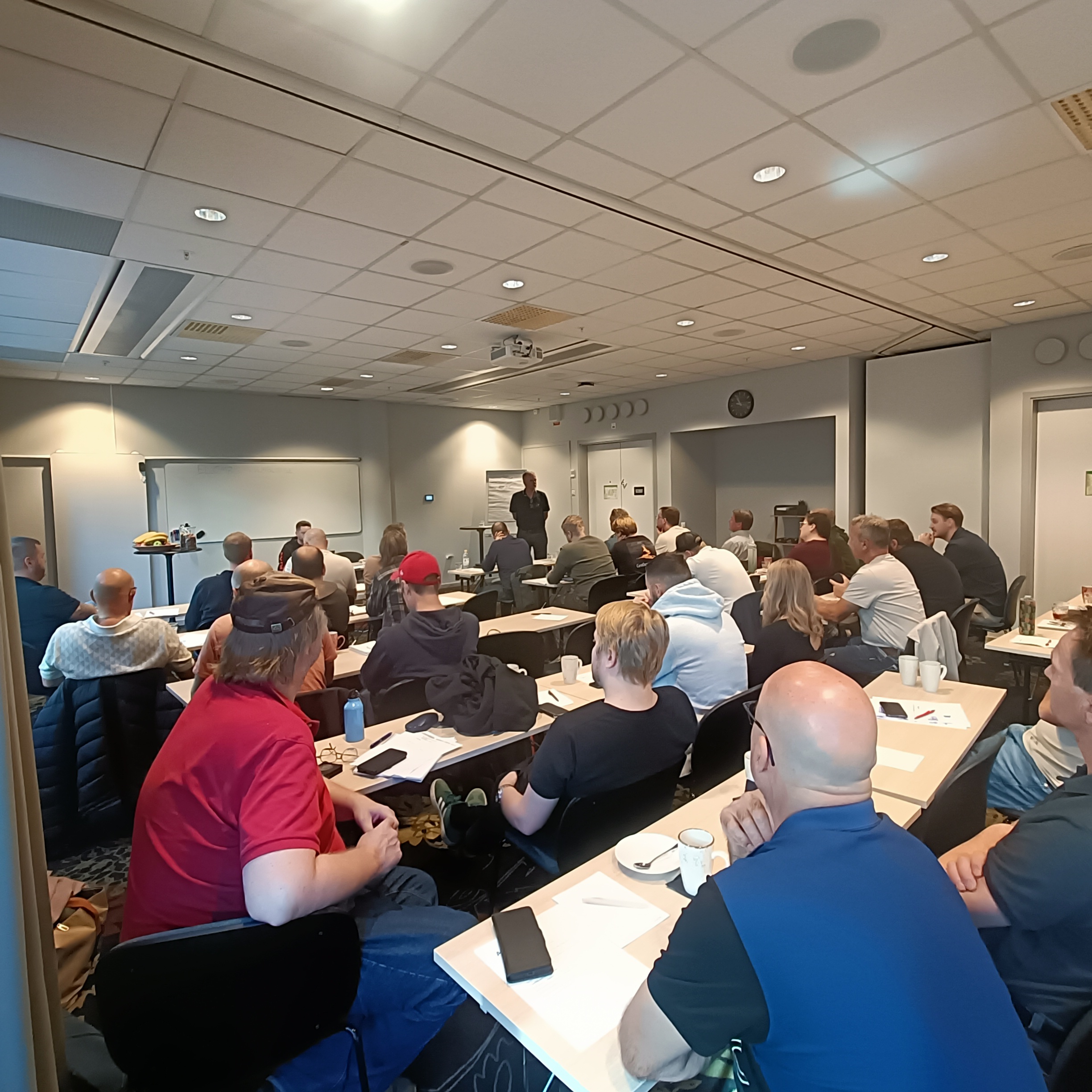 En grupp personer sitter vid bord i ett konferensrum och lyssnar på en talare som står vid en whiteboard och presentationsskärm.