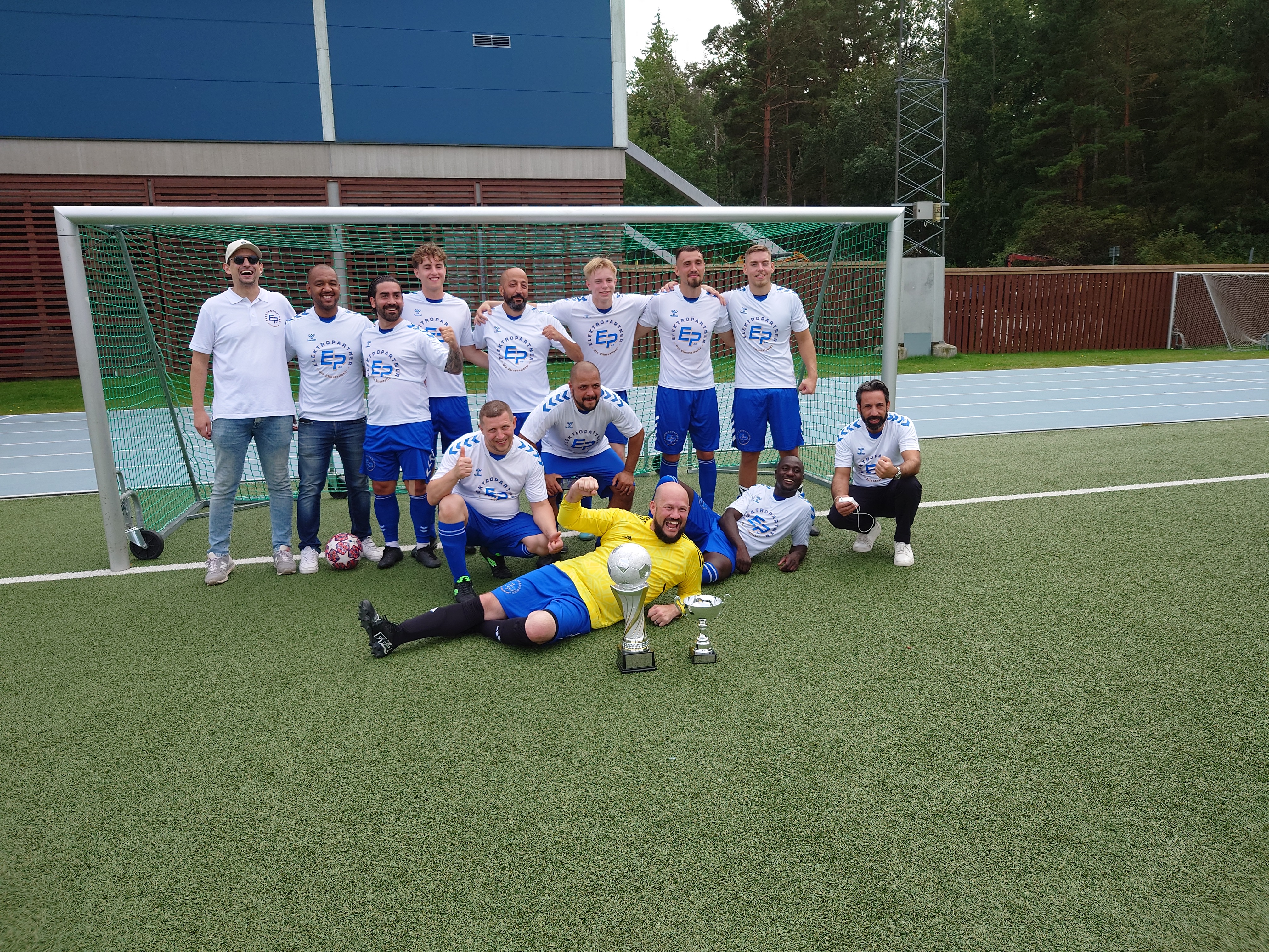 Ett fotbollslag poserar framför ett mål på en utomhusplan, några står och andra sitter, visar pokaler och en fotboll.