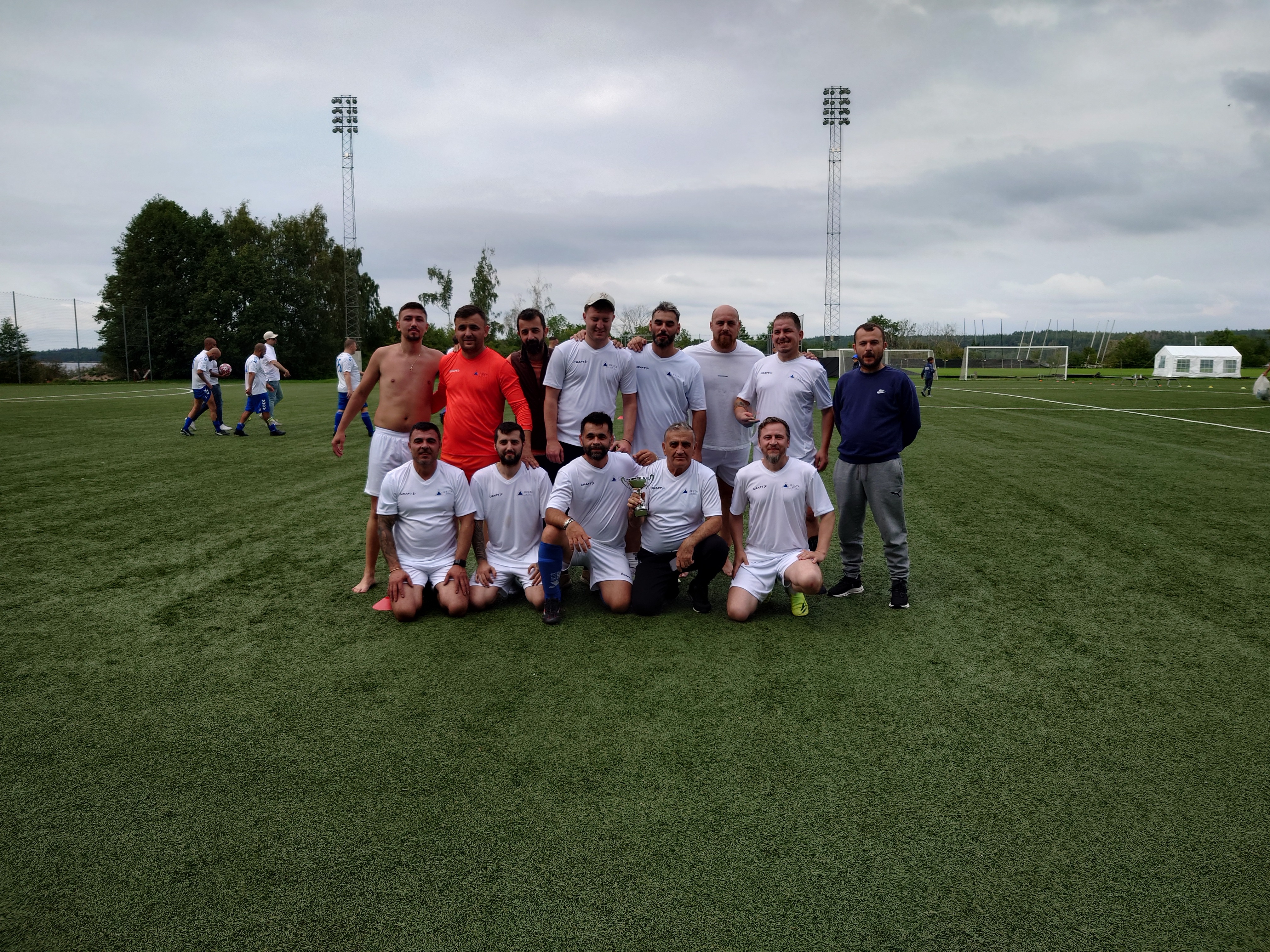 En grupp män poserar för ett foto på en fotbollsplan; de flesta har vita sporttröjor och shorts, målburar och lagkamrater syns i bakgrunden.