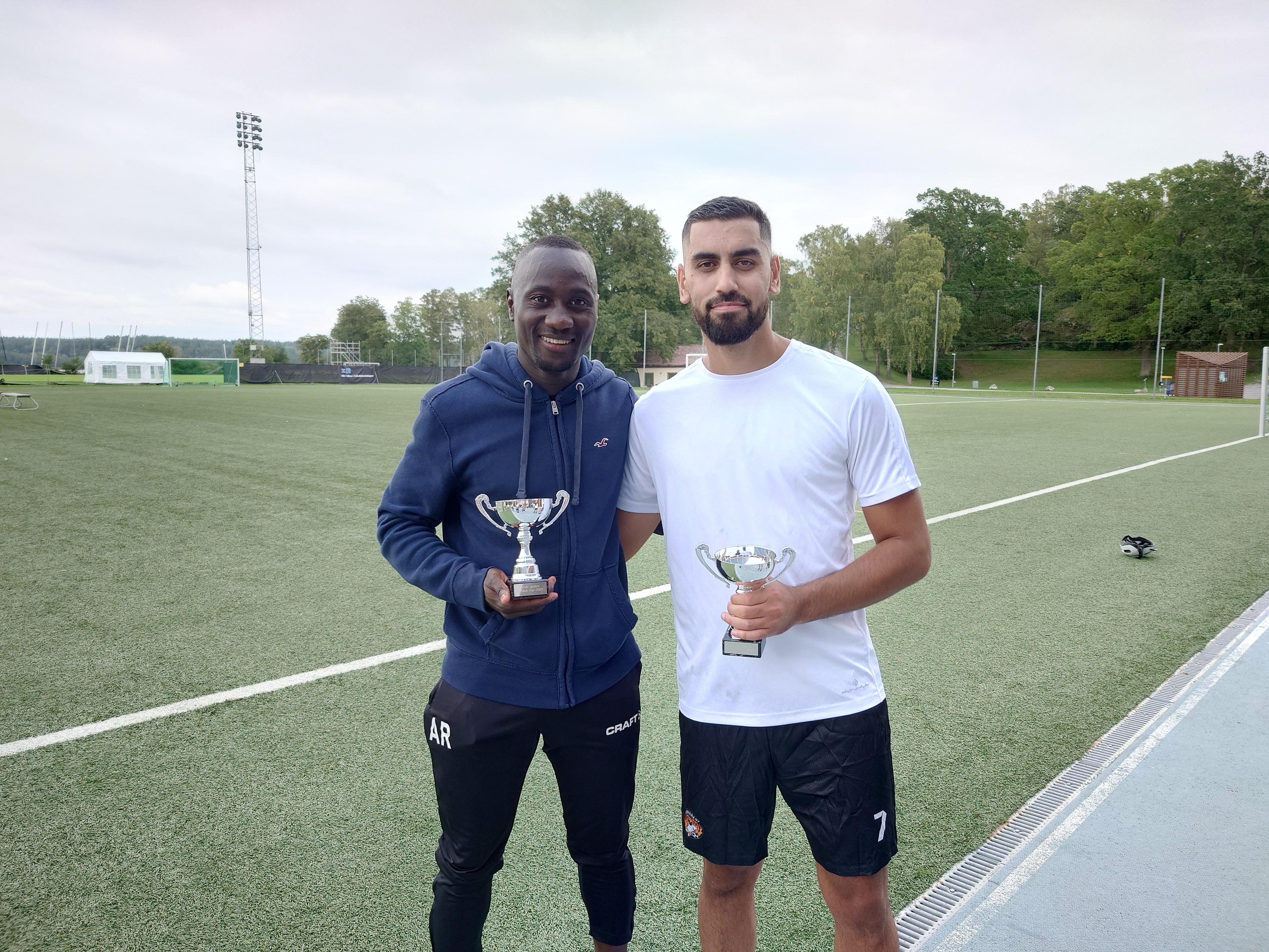 Två män står på en fotbollsplan, båda håller i en pokal och ler mot kameran.