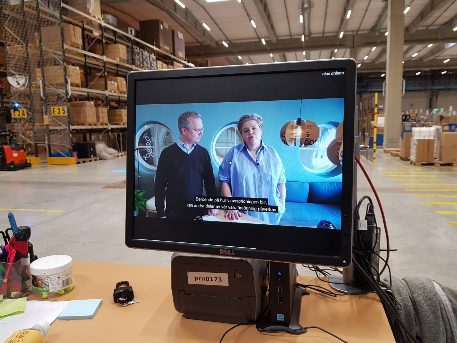 En datorskärm på ett skrivbord i ett lager visar ett tv-program med två personer och svenska undertexter, omgiven av kontors- och lagerutrustning.