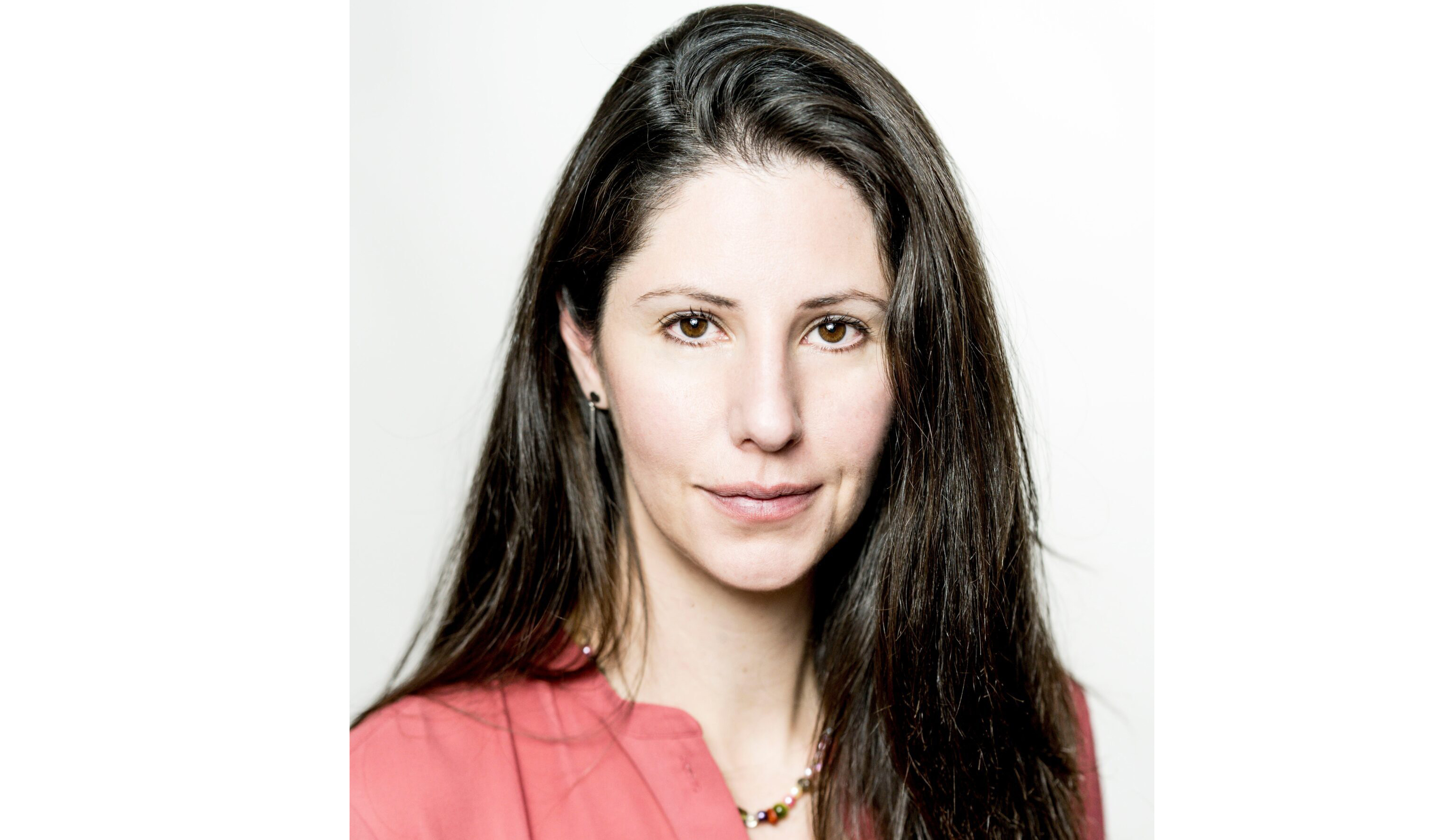 En kvinna med långt brunt hår och rosa topp tittar in i kameran mot en vit bakgrund.