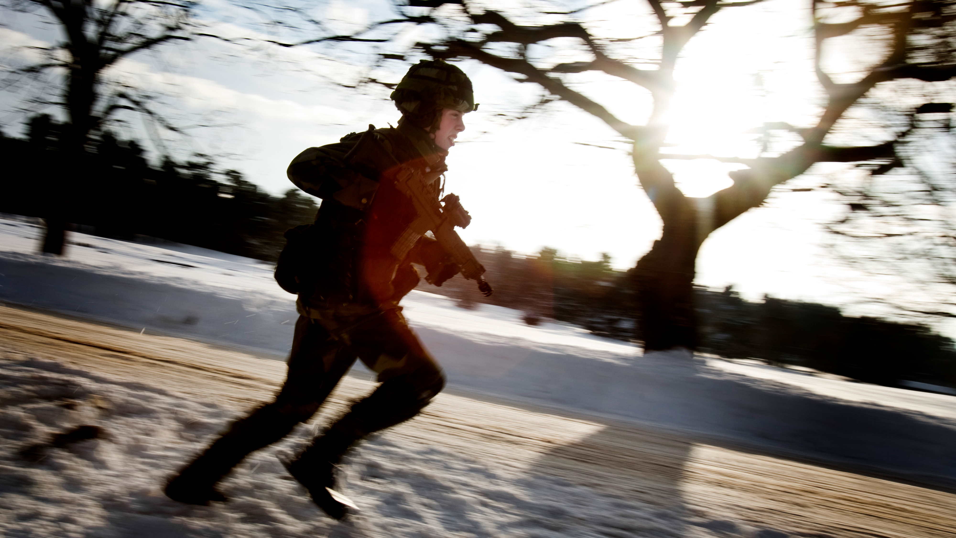 En soldat i uniform springer genom ett snötäckt landskap med ett gevär, framför solen och nakna träd.