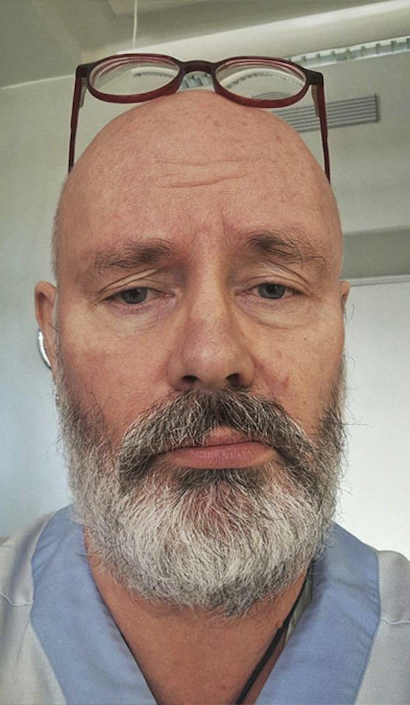 En medelålders man med rakat huvud, grått skägg och blå arbetskläder tittar mot kameran. Glasögon ligger på huvudet.