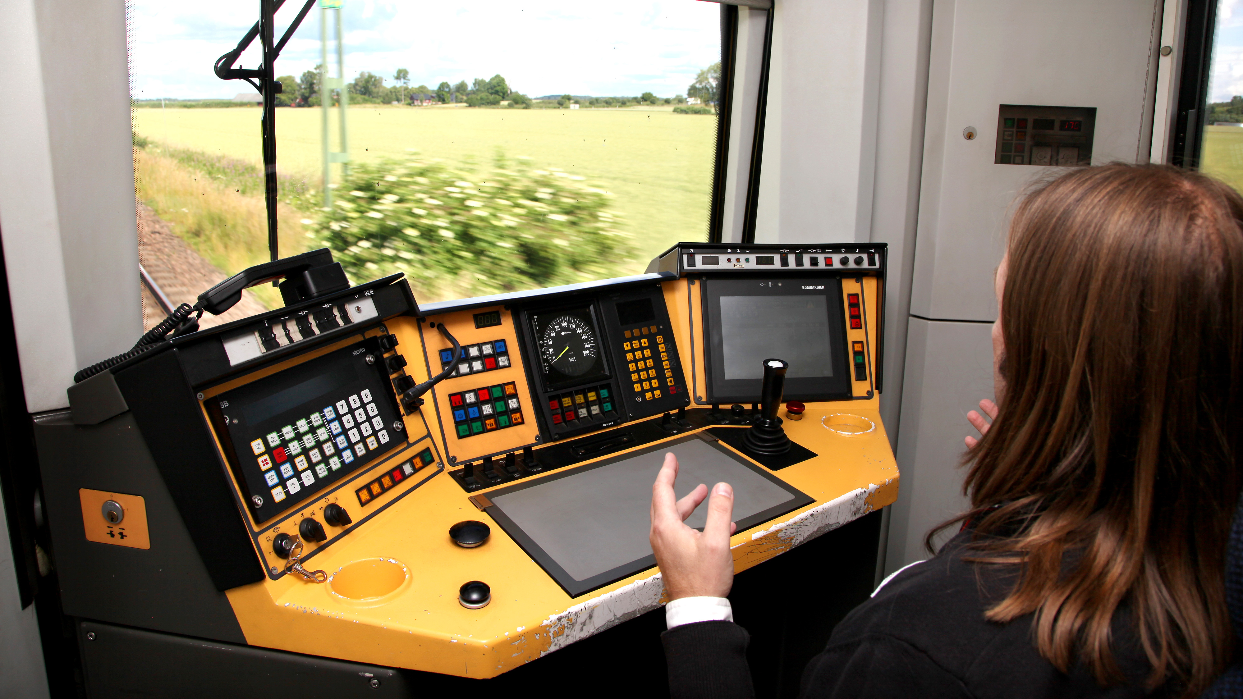 Person använder ett tågkontrollbord med flera skärmar, knappar och spakar och tittar ut genom fönstret mot landskapet.