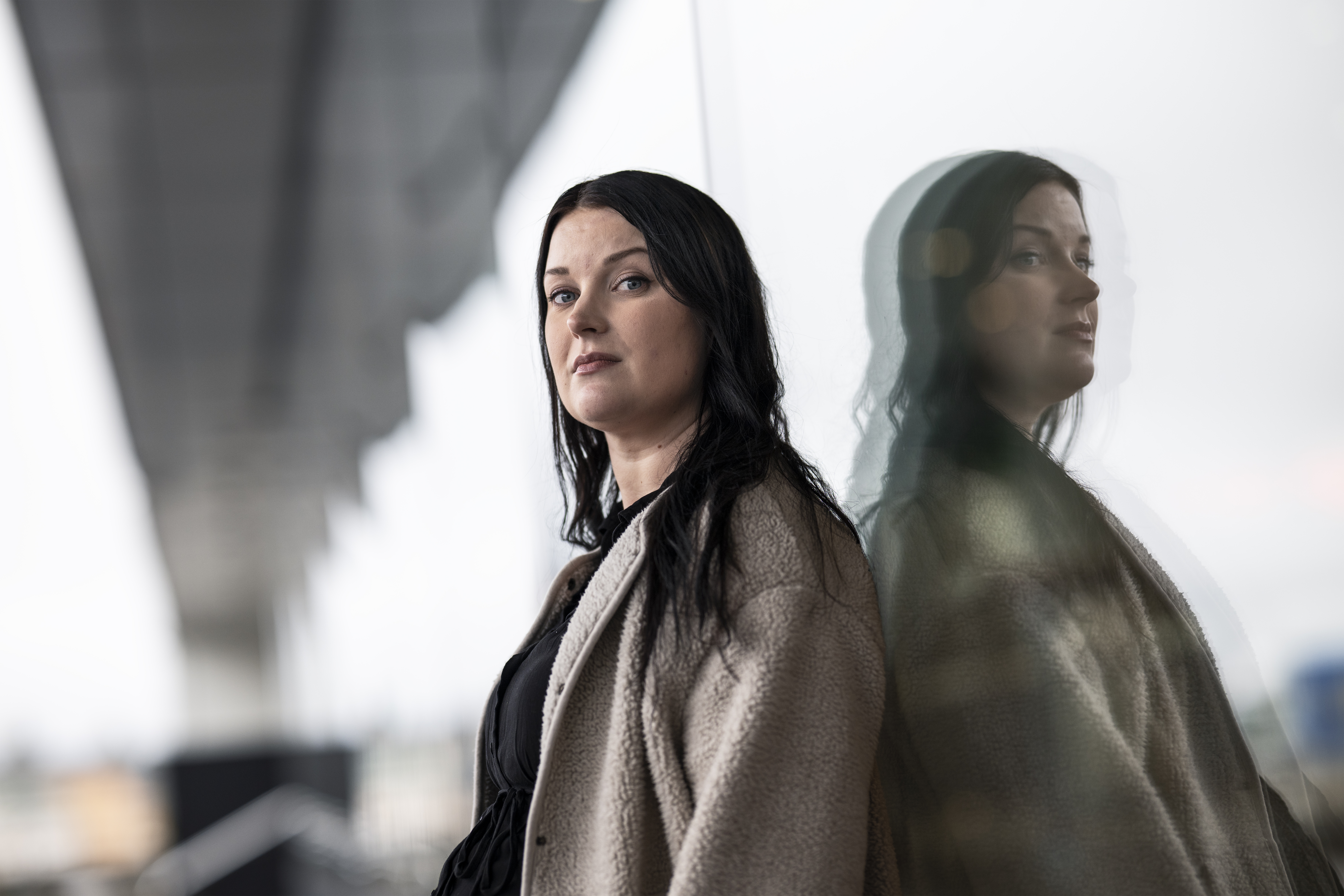 Maja Jansson med mörkt hår står inomhus och lutar sig mot en glasvägg där hennes spegelbild syns bredvid henne.