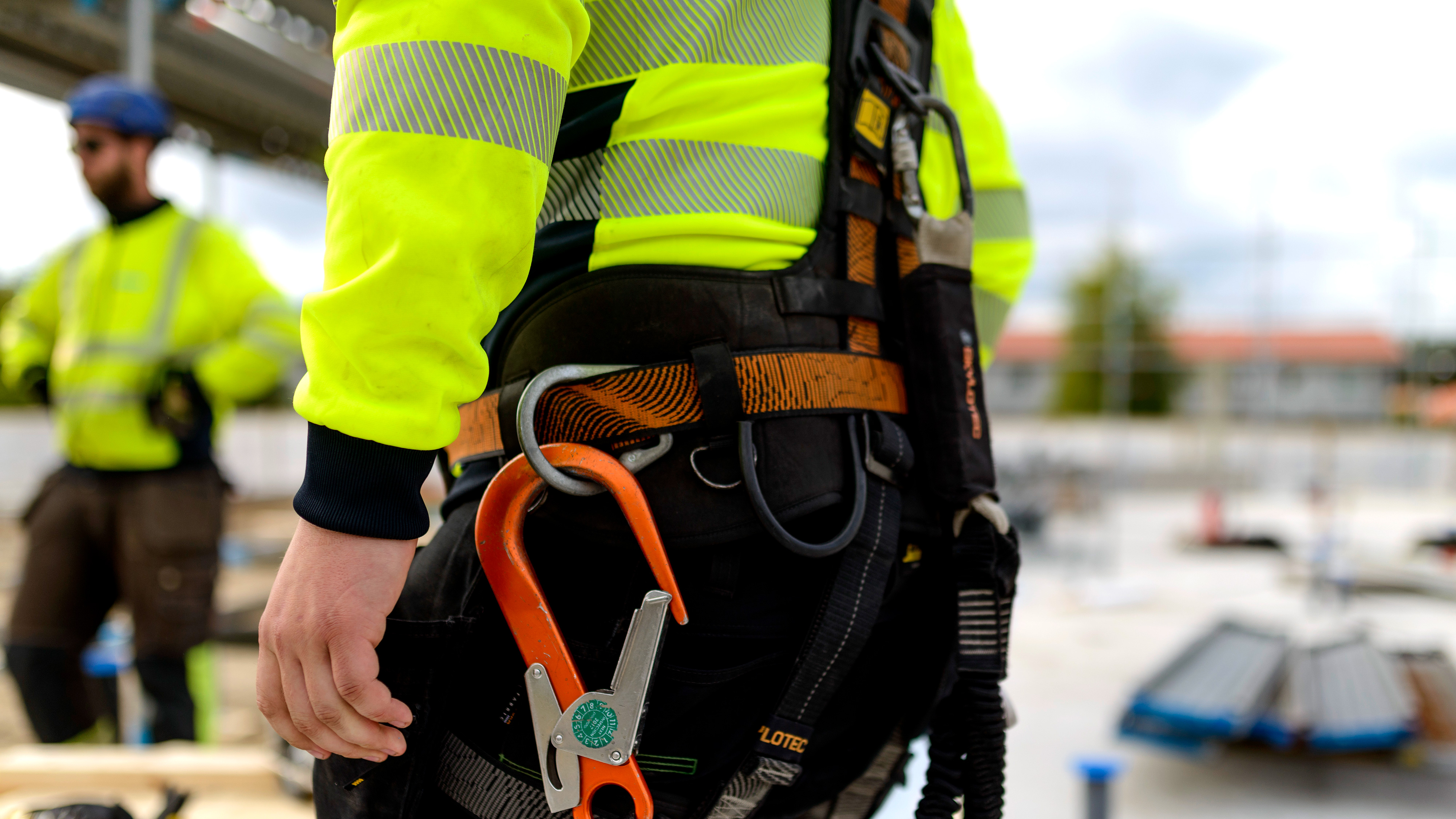 En byggnadsarbetare med säkerhetssele och reflexjacka står på en arbetsplats med utrustning i bältet. En annan syns bakom.