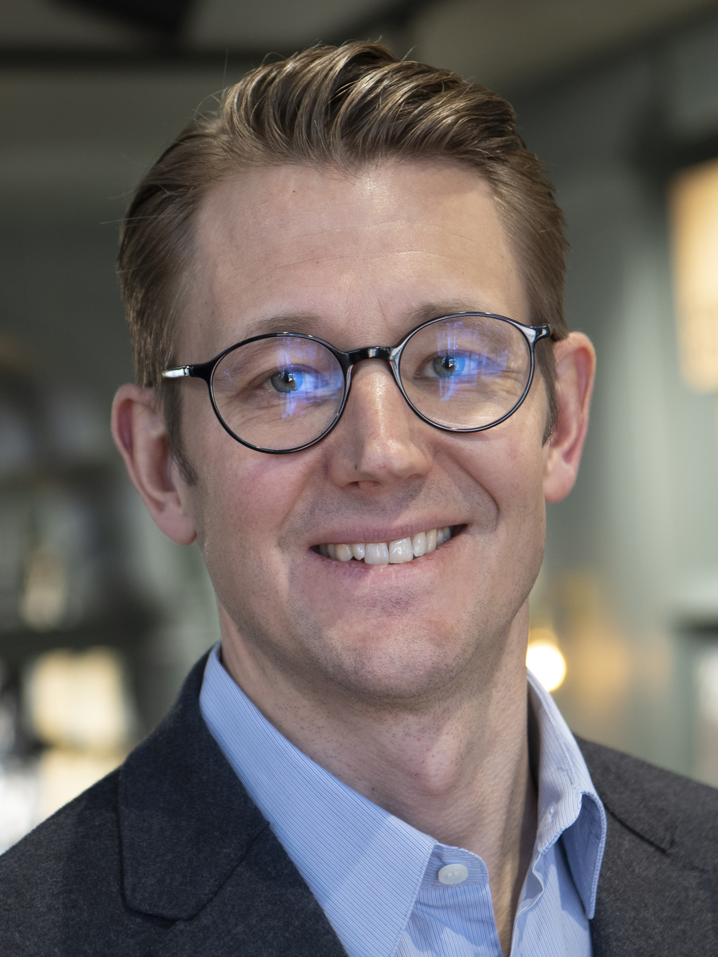 Kristoffer Tonström med kort brunt hår och glasögon ler. Han har på sig en blå skjorta och en mörk kavaj.