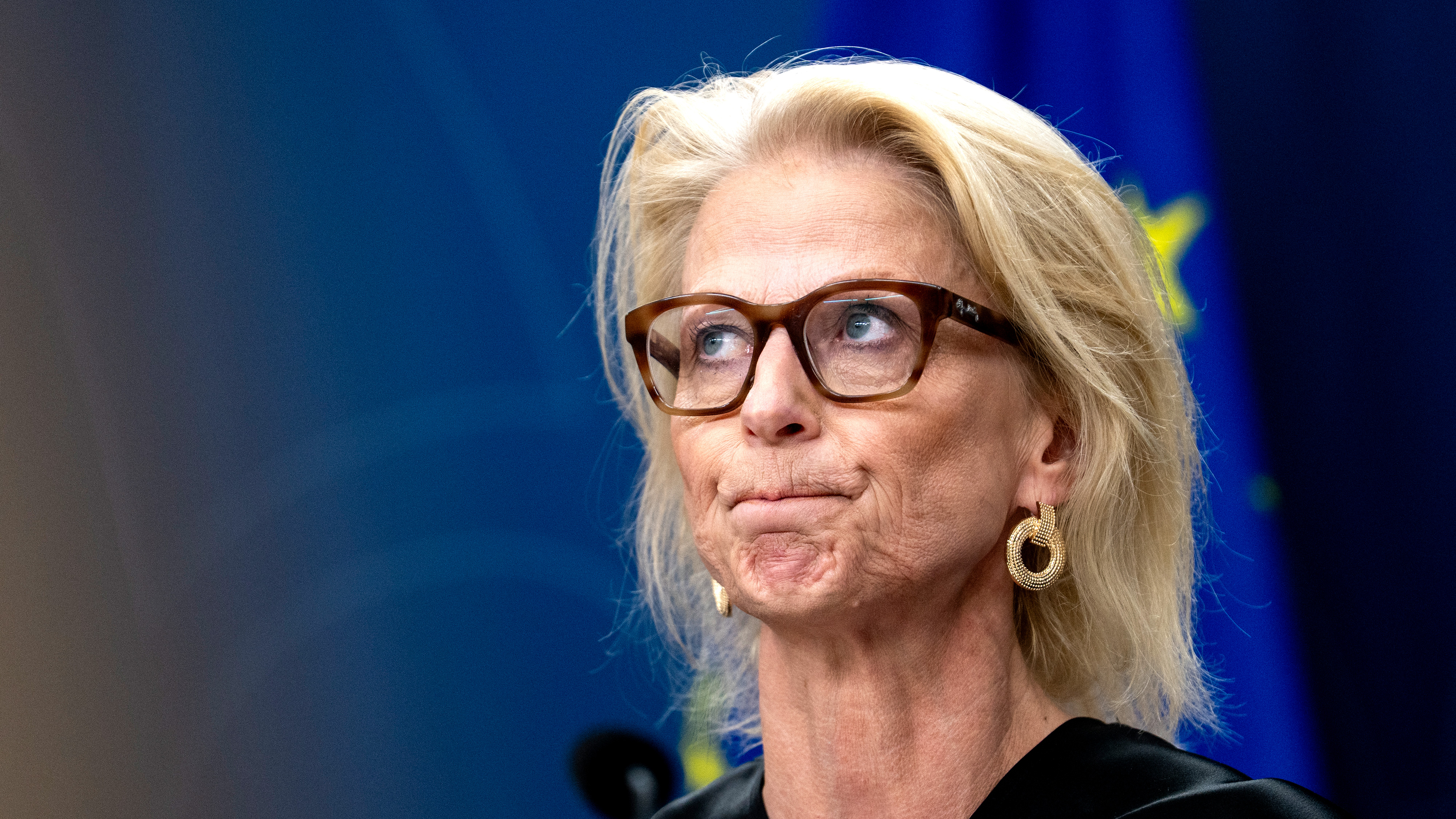 En kvinna med blont hår och glasögon står framför en blå bakgrund med en del av en gul stjärna, har örhängen och svart topp.