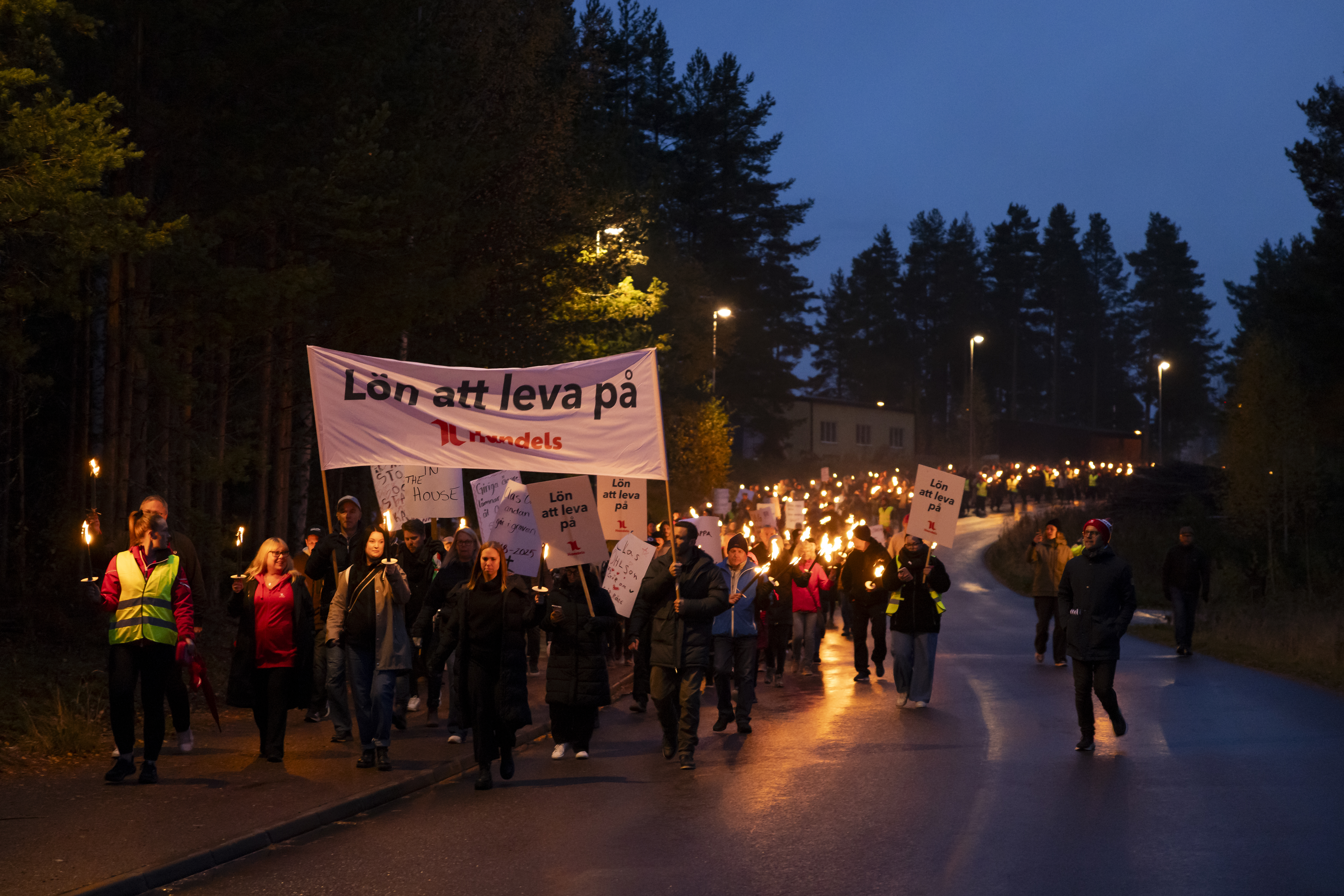 En grupp personer går på en väg med facklor och skyltar, bland annat ett stort plakat med texten "Lön att leva på".