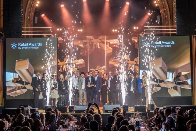 En grupp personer står på en scen och håller pokaler vid Retail Awards, med fyrverkerier och stora skärmar i bakgrunden, publik tittar på.
