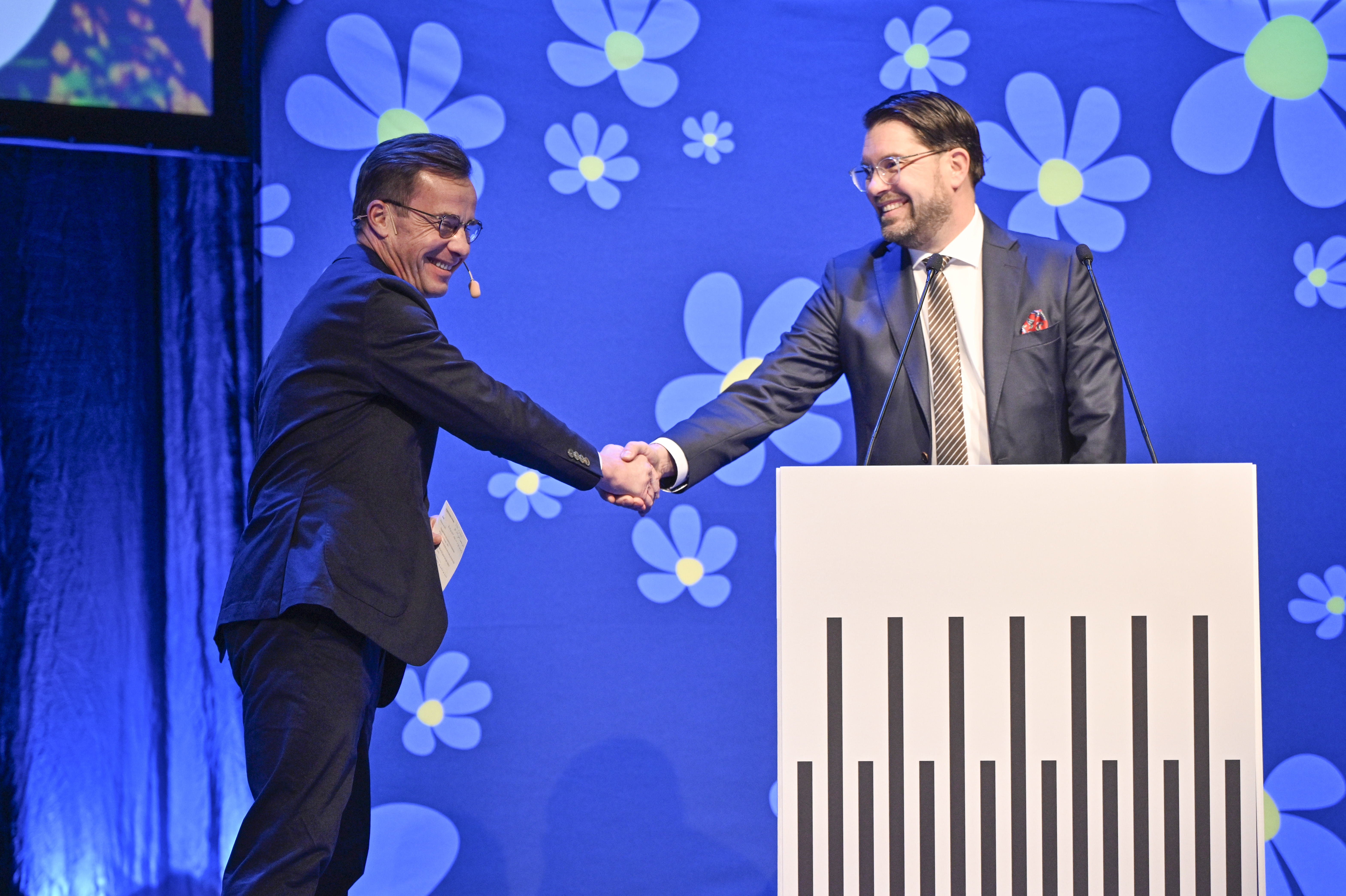 Ulf Kristersson och Jimmie Åkesson tar i hand på en scen med blå bakgrund och vita samt blå blommor.