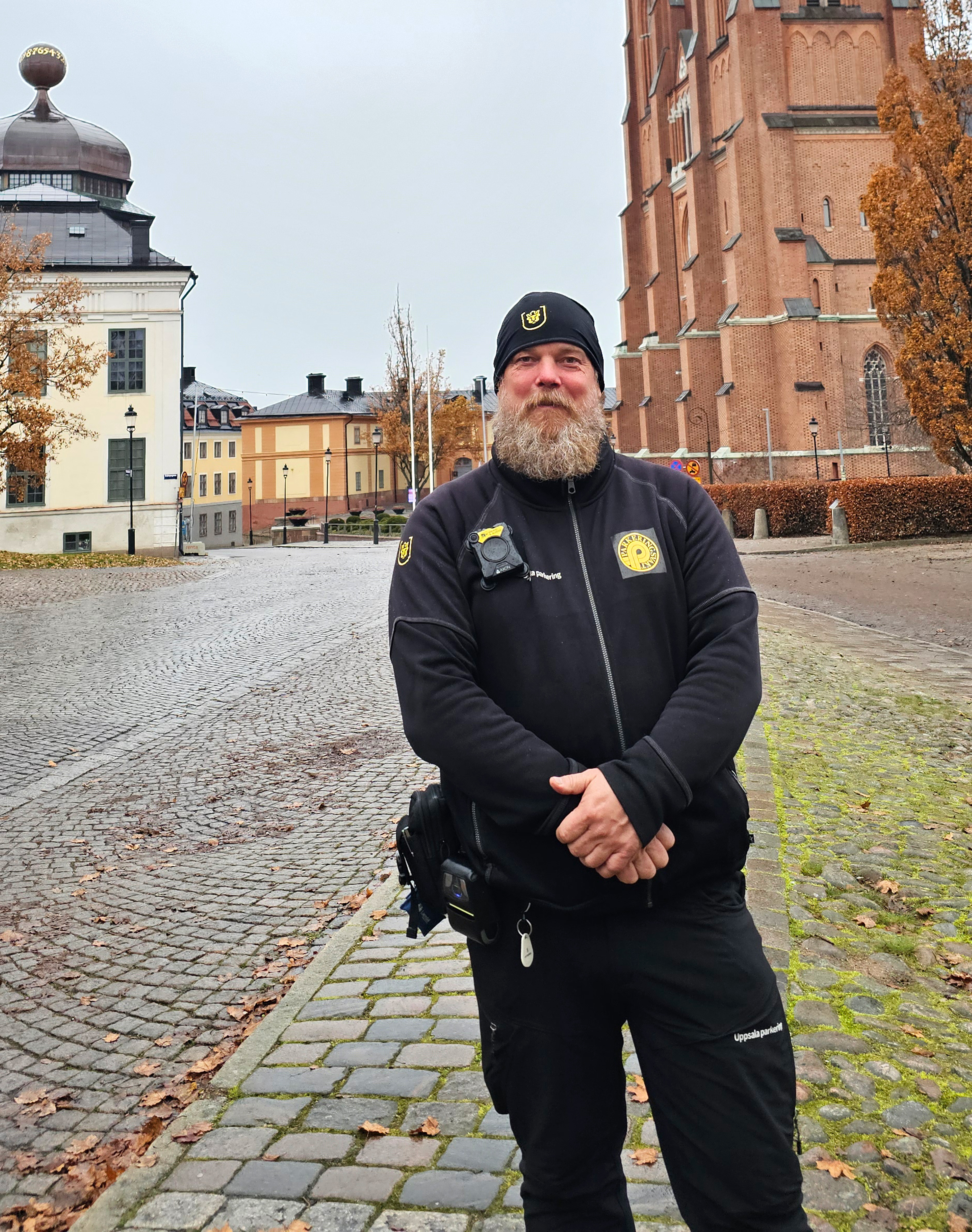 En skäggig man i svart uniform och hatt står på en gatstenbelagd gata framför äldre byggnader och träd med höstlöv.