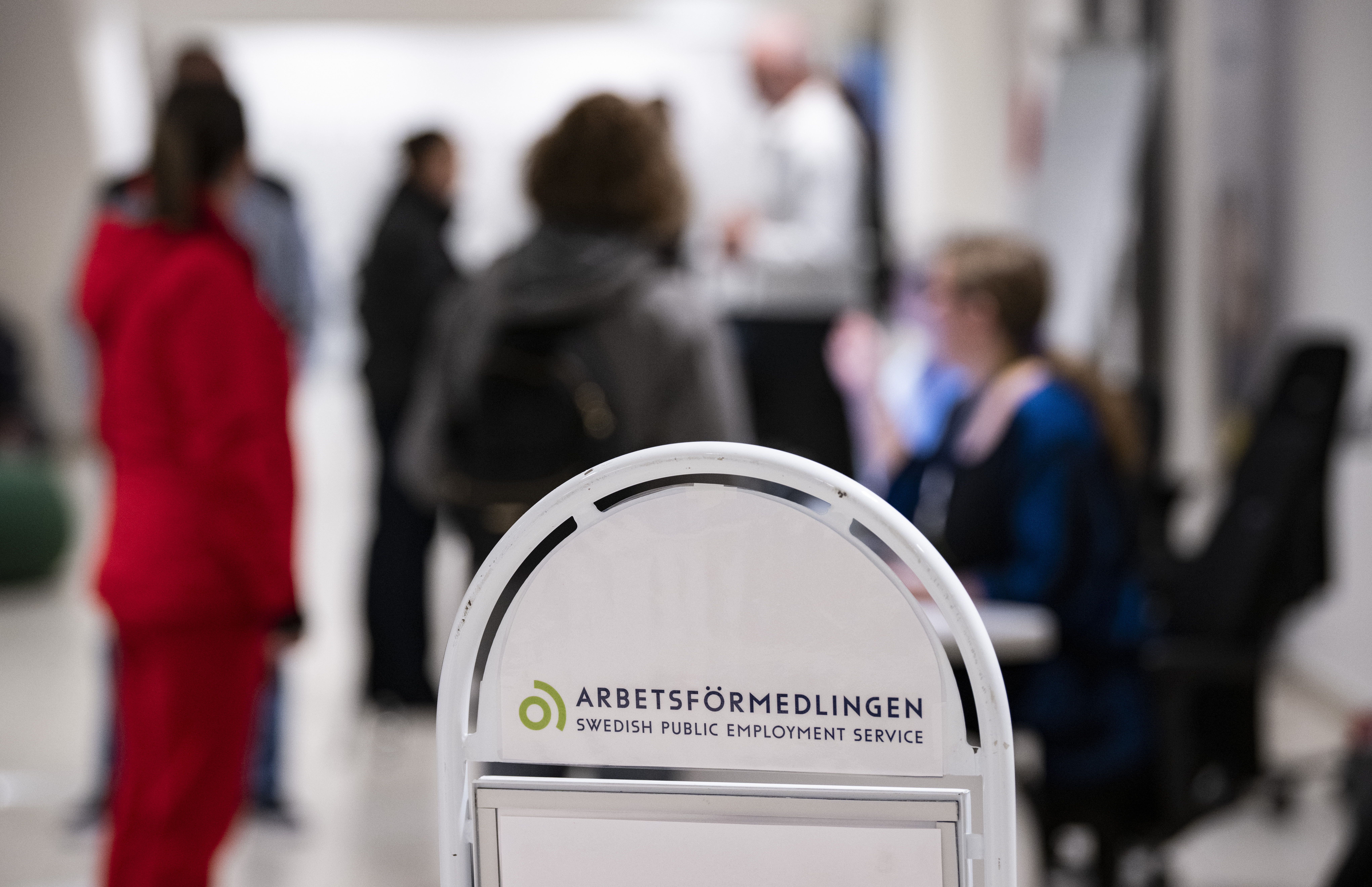 Skylt för Arbetsförmedlingen i förgrunden, flera personer står samlade och samtalar i bakgrunden på ett kontor.