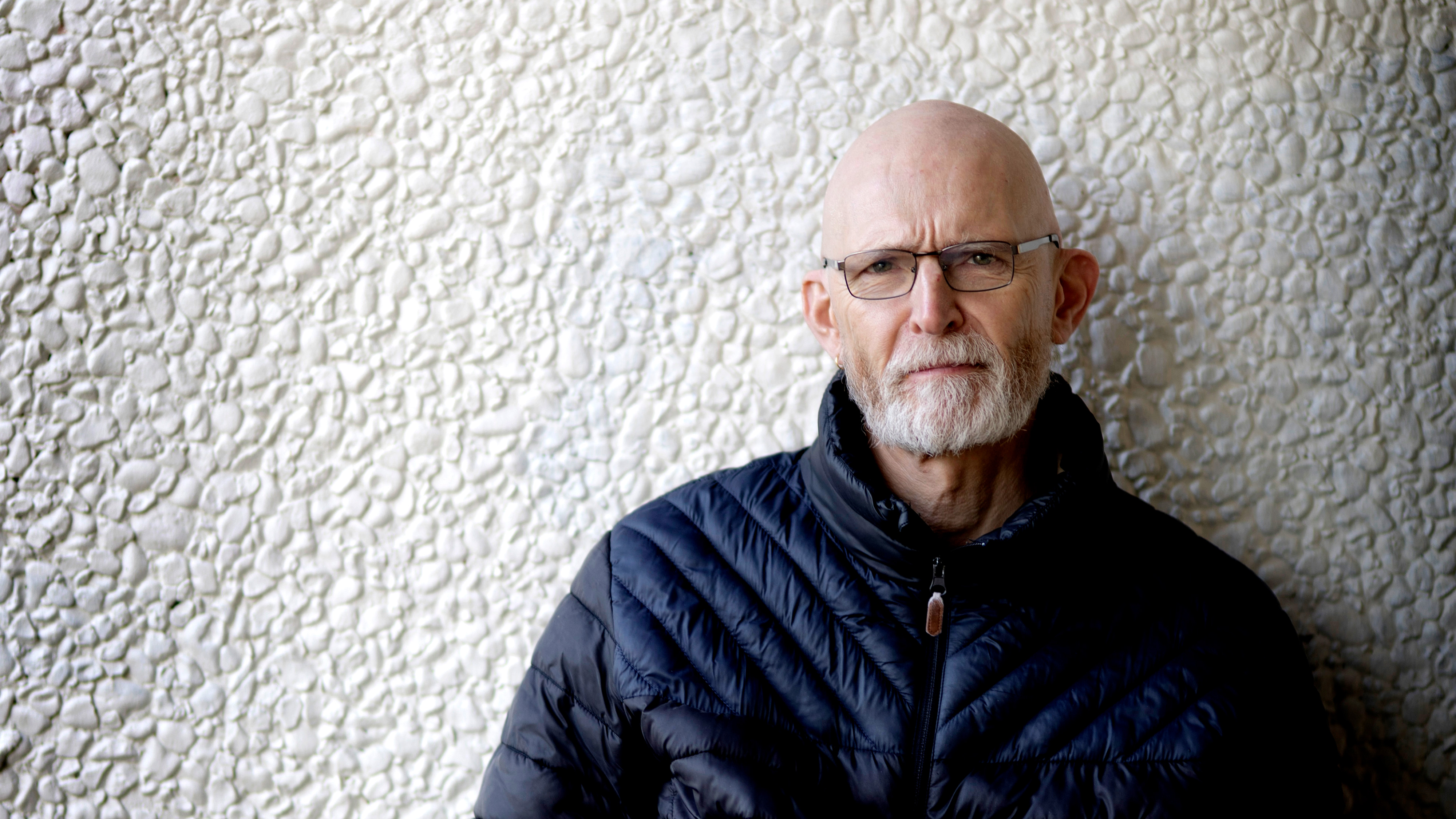 Skallig man med glasögon och vitt skägg, i mörk jacka, står framför en vit vägg.