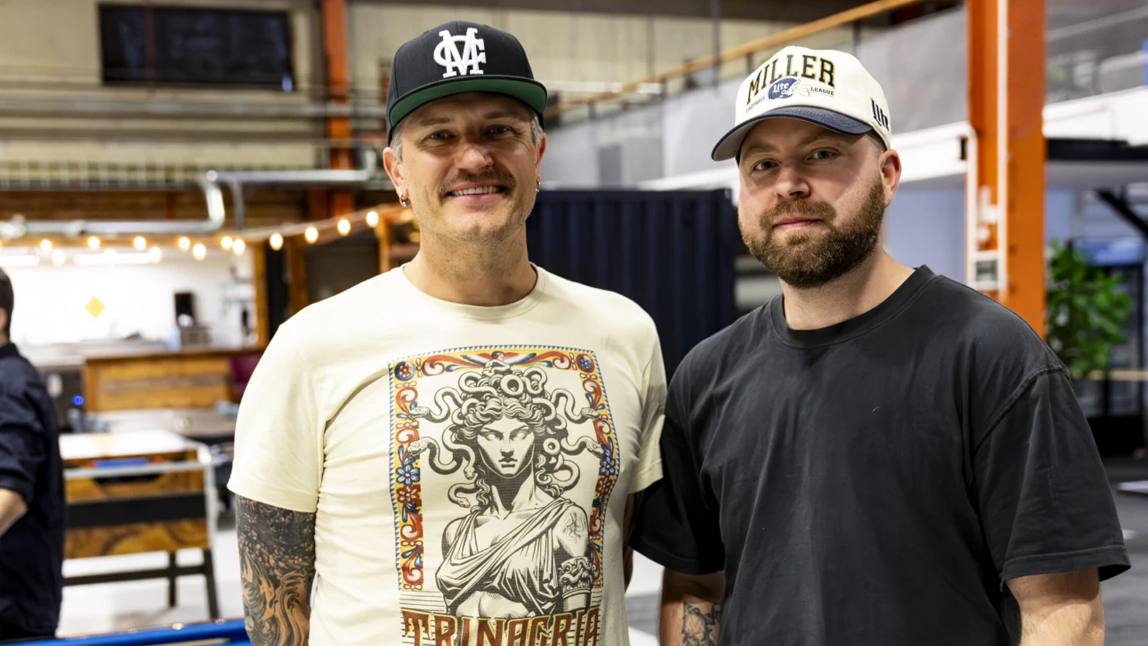 Två män står bredvid varandra inomhus, båda har keps. En har en t-shirt med tryck, den andra en enfärgad mörk tröja. Bakgrunden visar ett öppet kontorsutrymme.