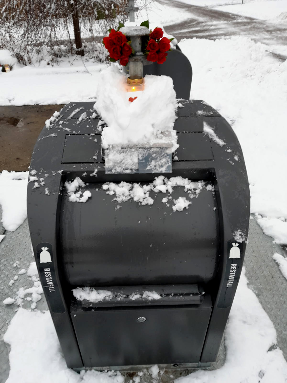 En svart soptunna står i snön med ett ljus, röda rosor och en hjärtdekoration ovanpå.