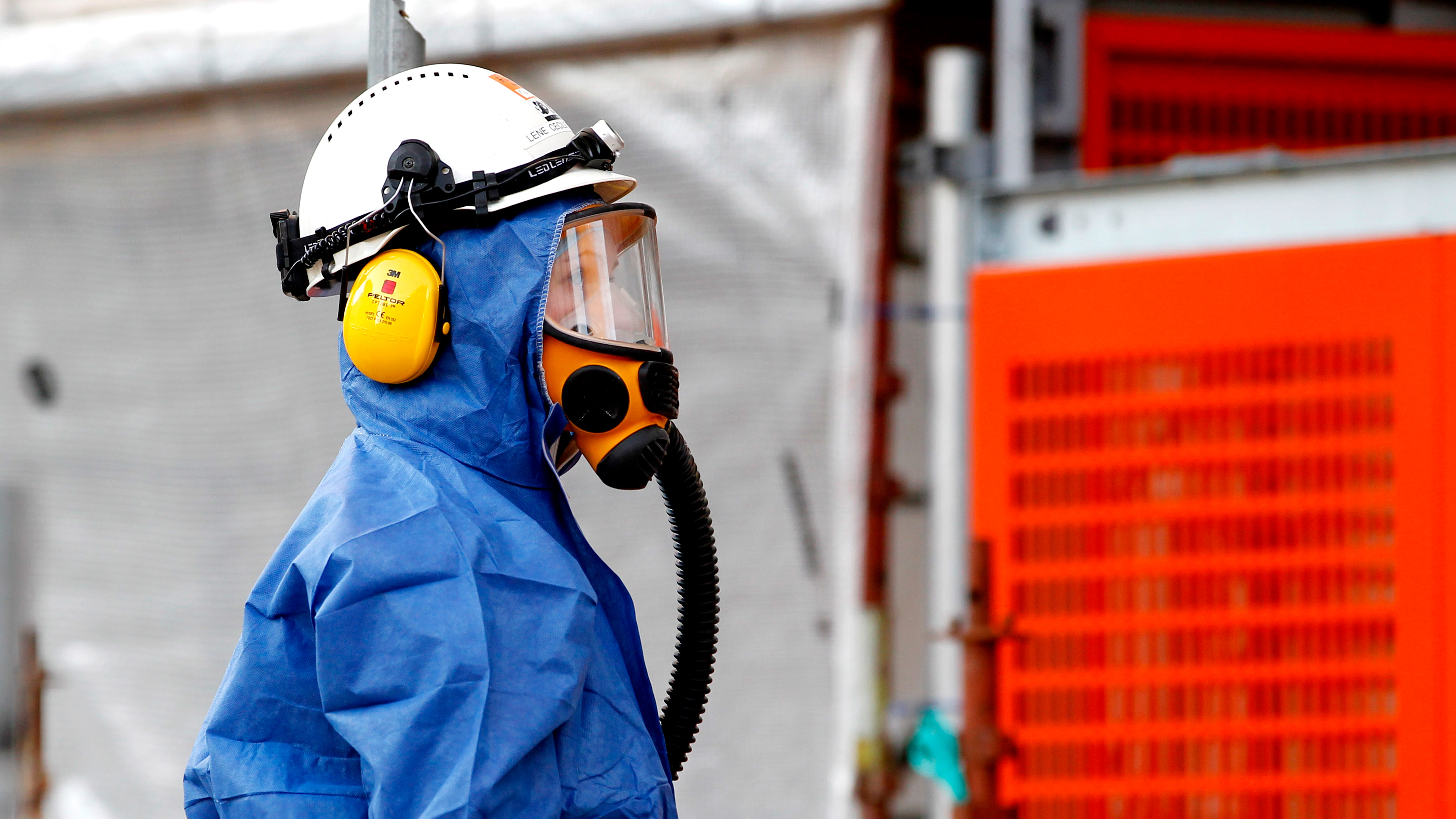 Person i blå skyddsdräkt, hjälm, ansiktsmask med andningsskydd och hörselskydd står i ett industriområde.