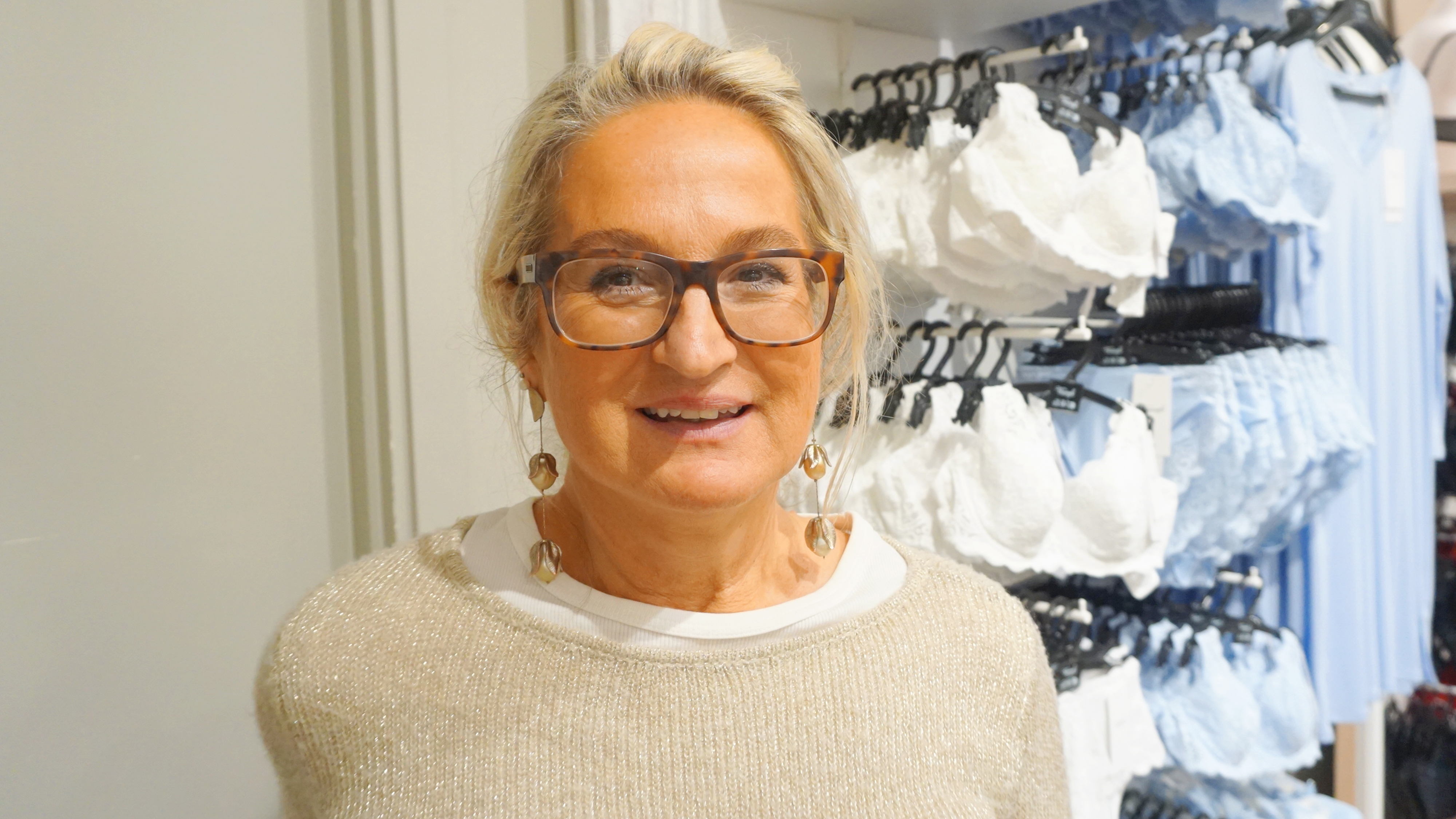 En kvinna med glasögon och örhängen står i en butiksgång med vita och ljusblå behåar på galgar i bakgrunden.
