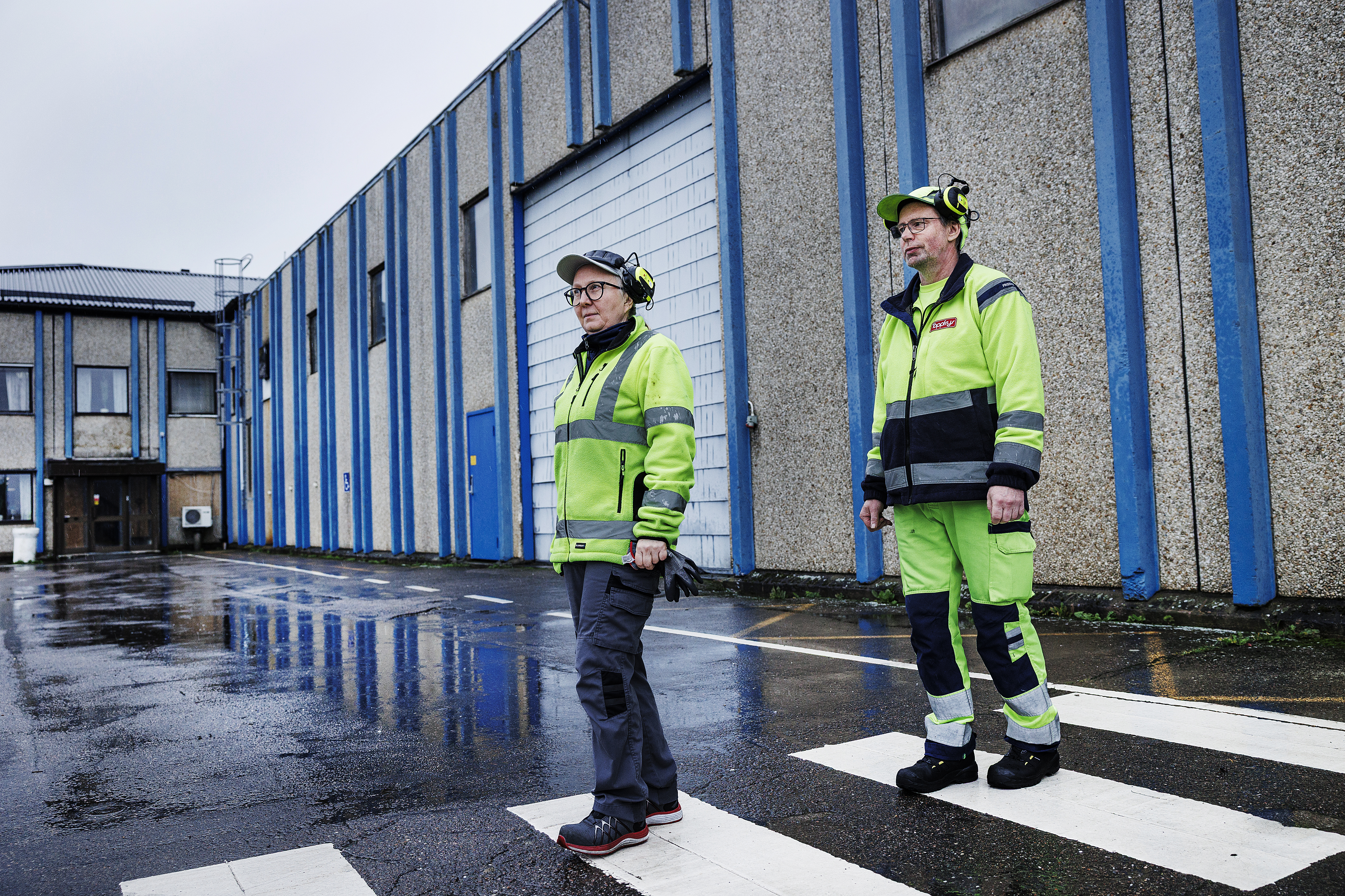 Två arbetare i varselkläder står på ett övergångsställe utanför en industribyggnad under en mulen och regnig dag.
