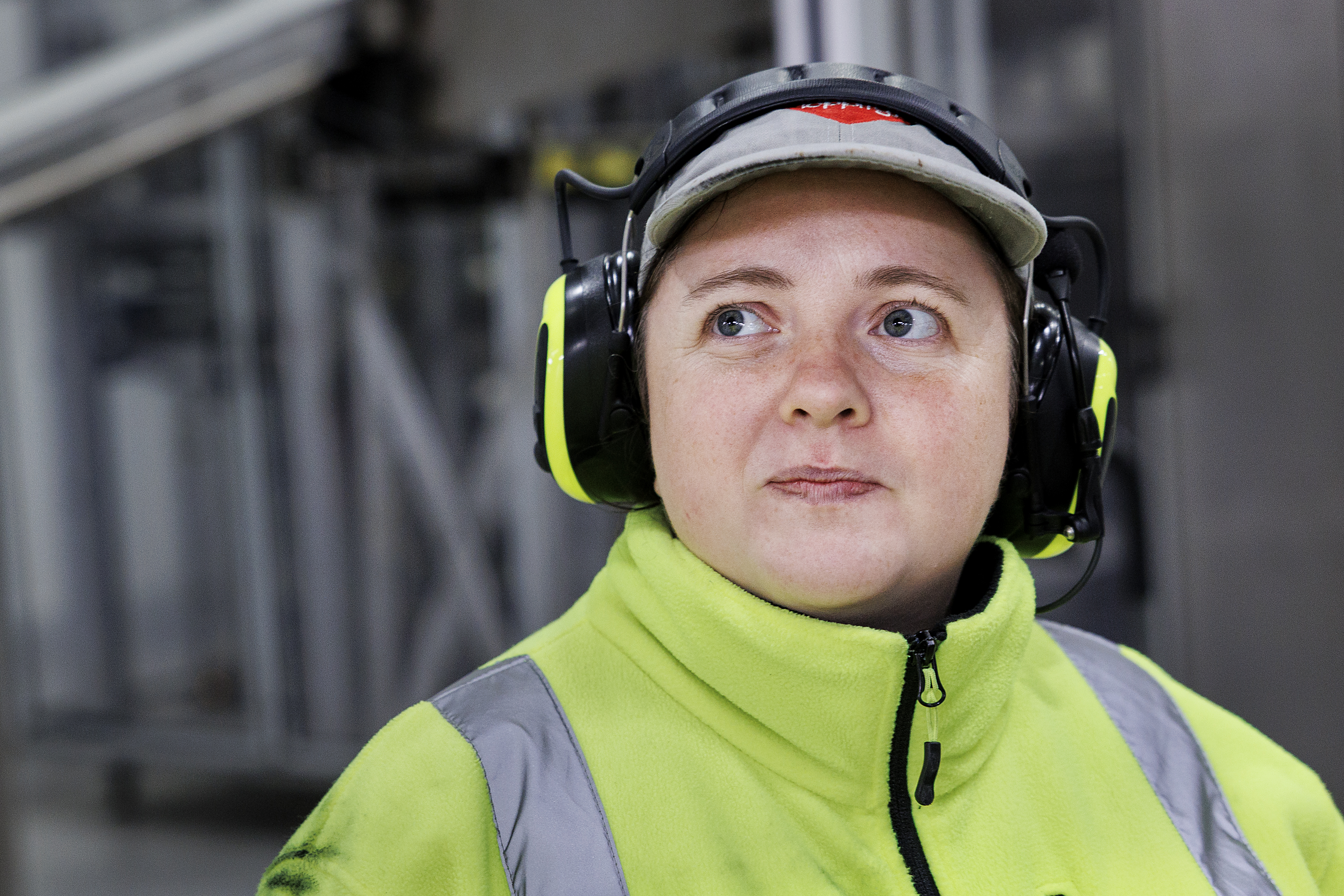 En person med varseljacka, keps och hörselskydd står inomhus och tittar lite åt sidan.