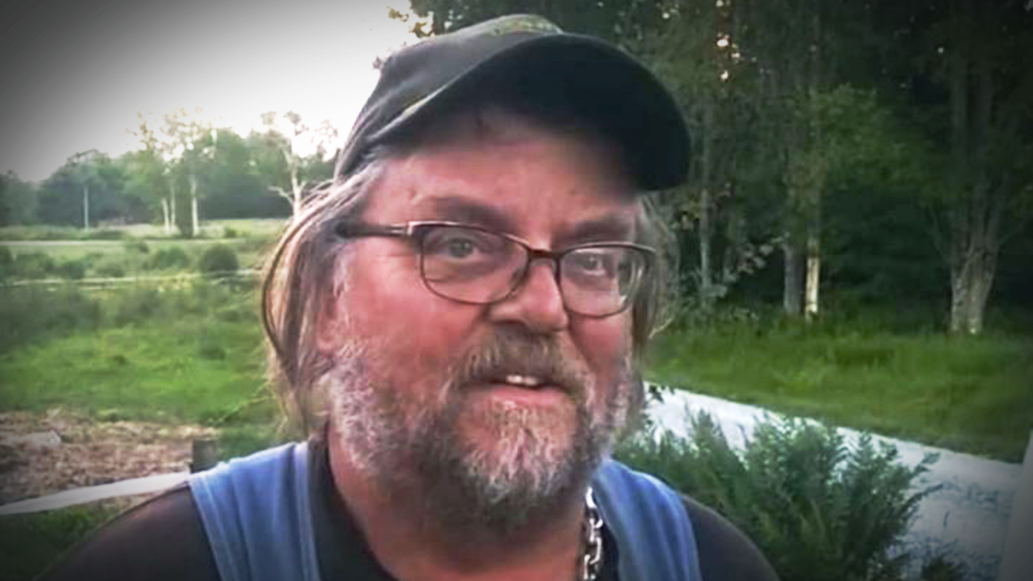 Lasse med glasögon, keps och skägg står utomhus framför växter, har på sig en svart tröja och kedjehalsband.