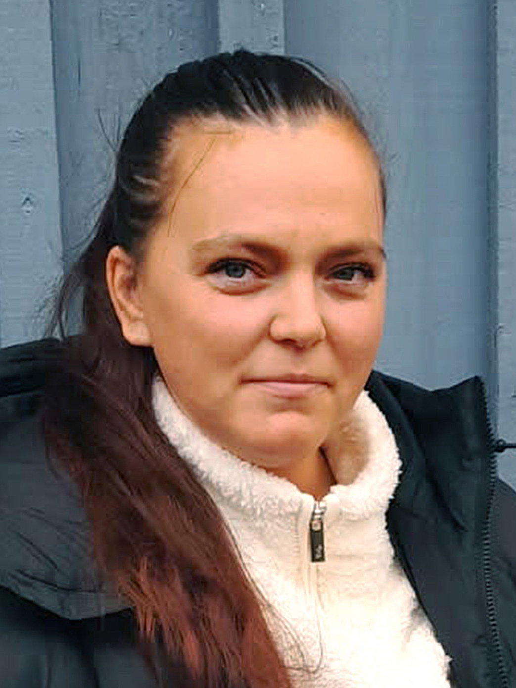 En kvinna med långt brunt hår uppsatt, har på sig en vit fleecejacka och en mörk jacka, står framför en blå trävägg.