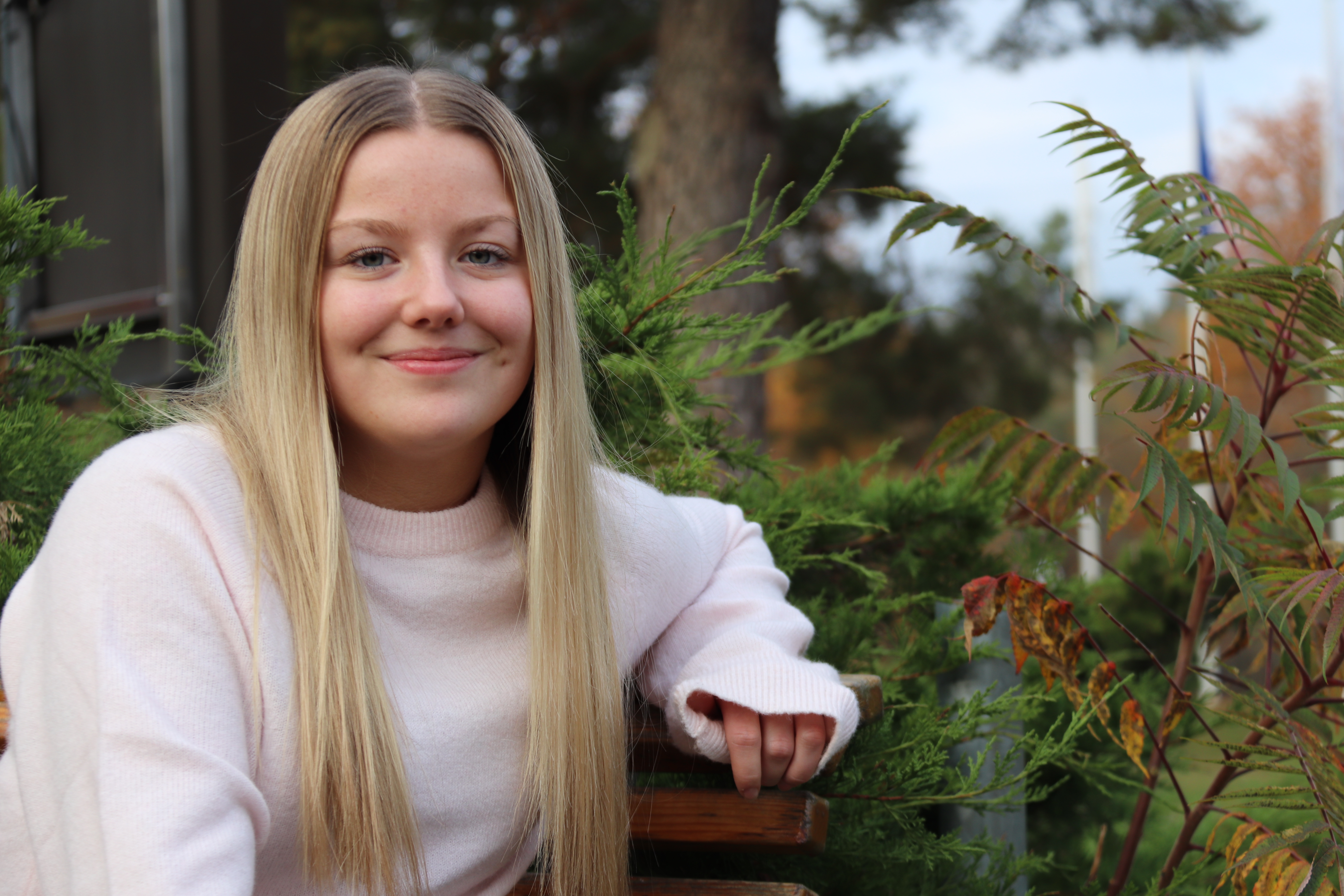 En ung kvinna med långt blont hår och en ljusrosa tröja sitter på en träbänk utomhus omgiven av grönska.