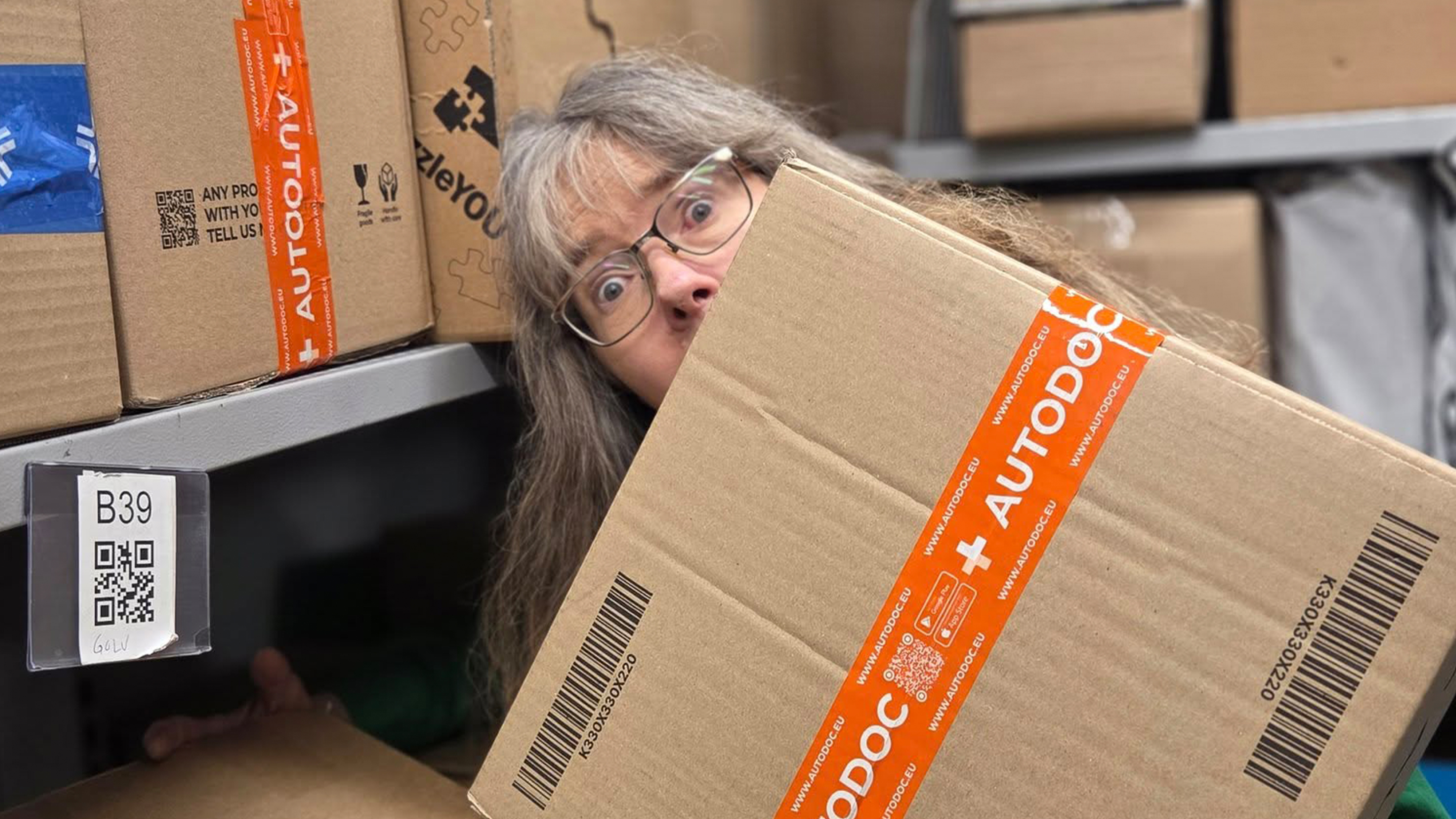 En person med långt hår och glasögon tittar fram bakom en kartong märkt "AUTODOC" i ett förråd med andra kartonger.