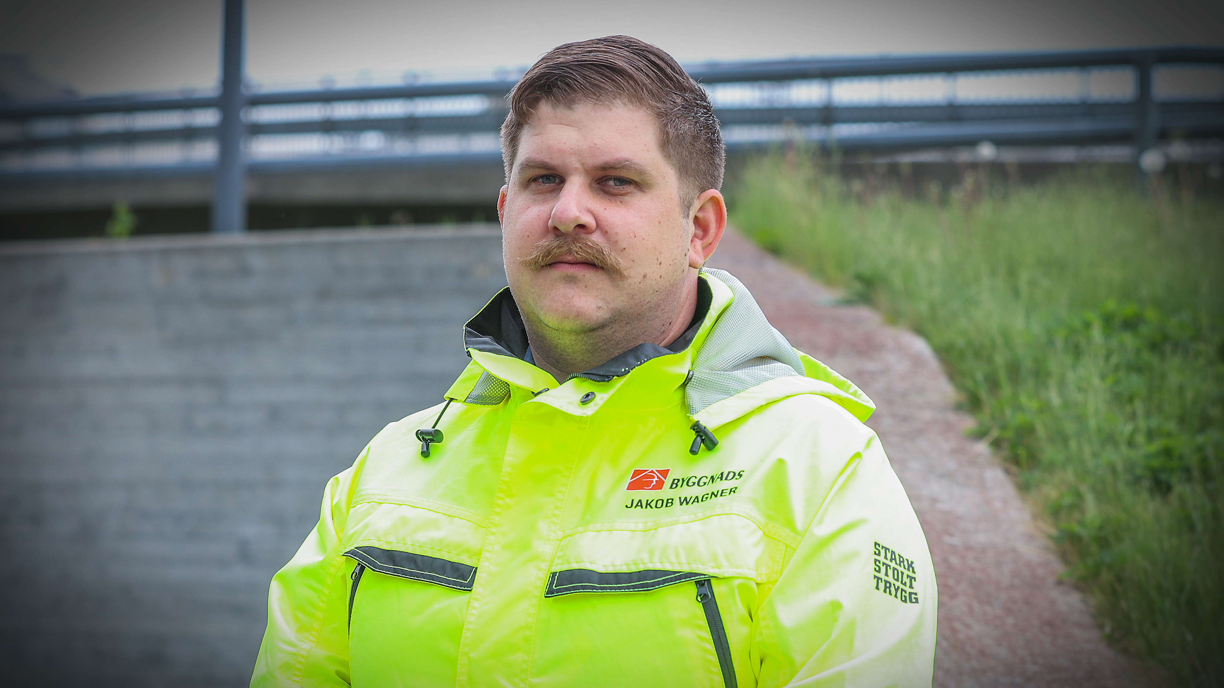 Jakob Wagner i gul arbetsjacka står utomhus nära en grässlänt och en betongvägg och tittar mot kameran.