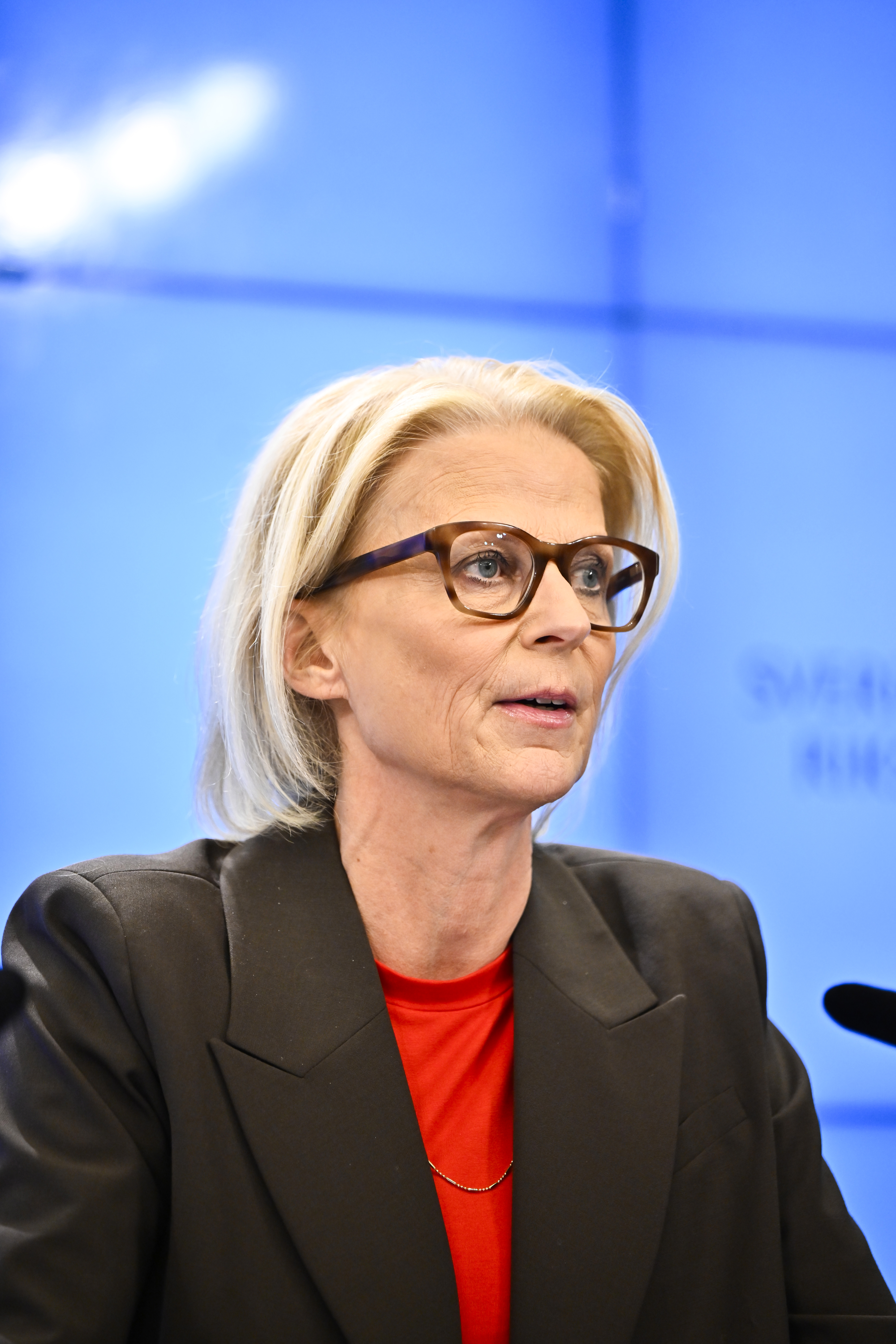 Finansminister Elisabeth Svantesson (M), iklädd svart kavaj och röd topp, talar vid ett evenemang med blå bakgrund.