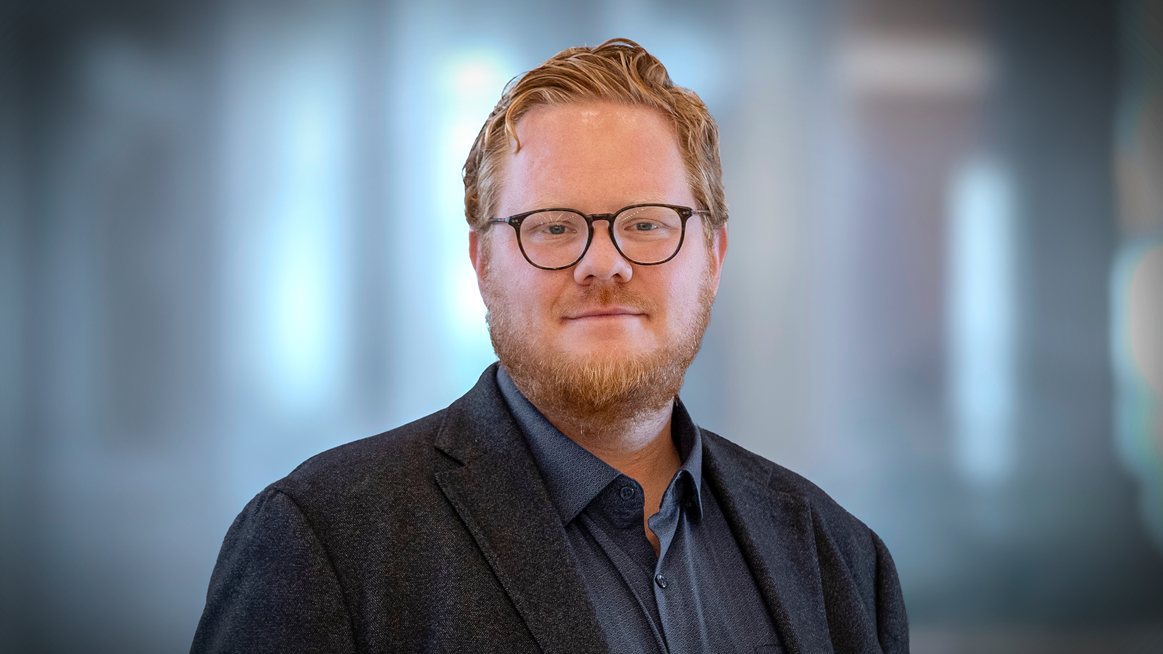 Emil Johansson med kort rött hår, glasögon och skägg har en mörk kavaj och blå skjorta, står framför en suddig inomhusmiljö.