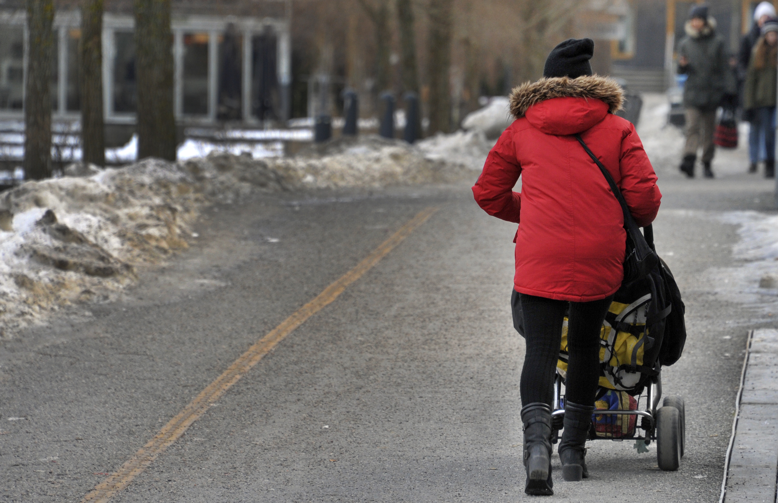 En person med röd jacka och svart hatt skjuter en barnvagn på en snötäckt gångväg med flera personer i bakgrunden.
