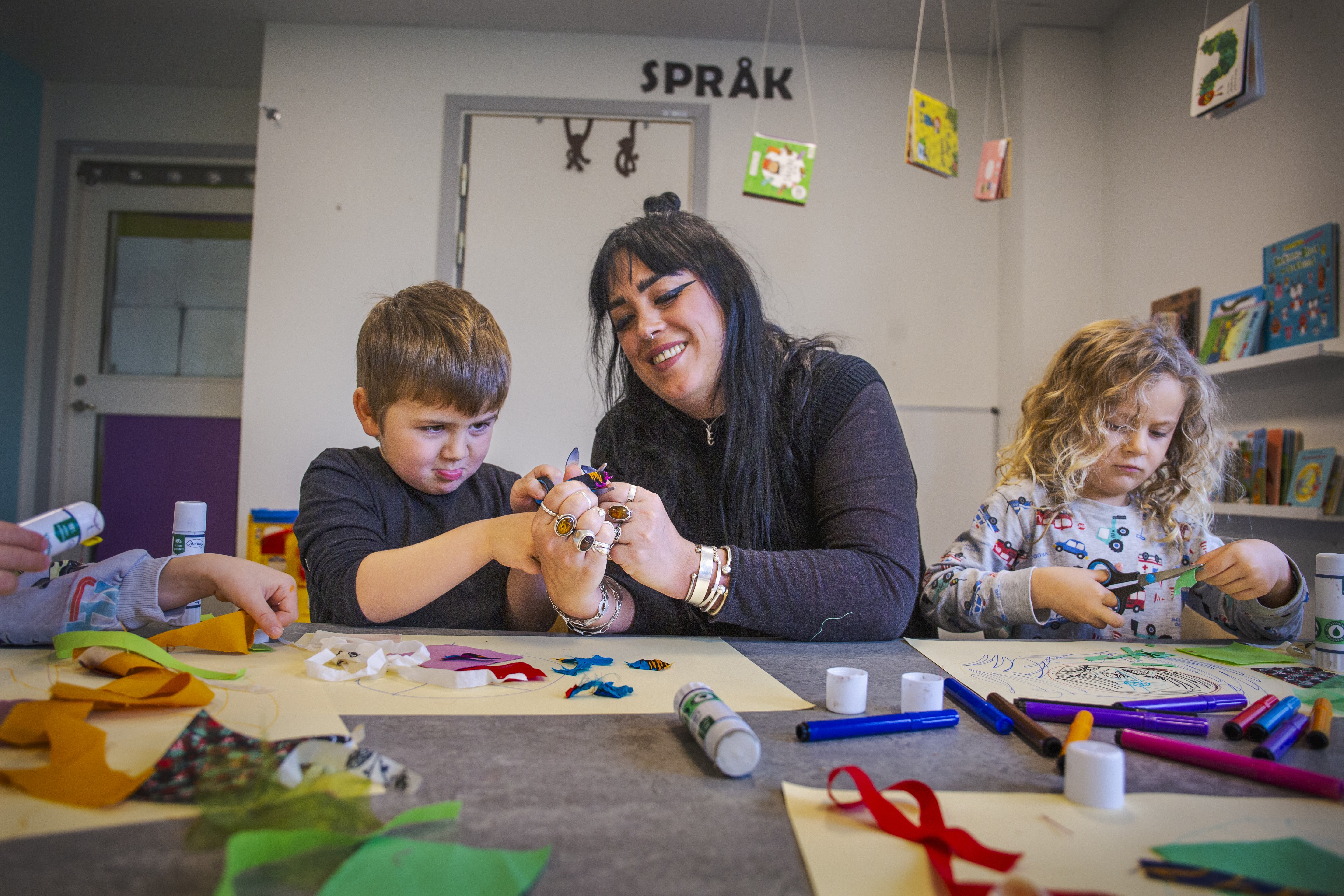 Myrna Vahlström hjälper ett barn att använda ett limstift medan ett annat barn klipper med en sax.