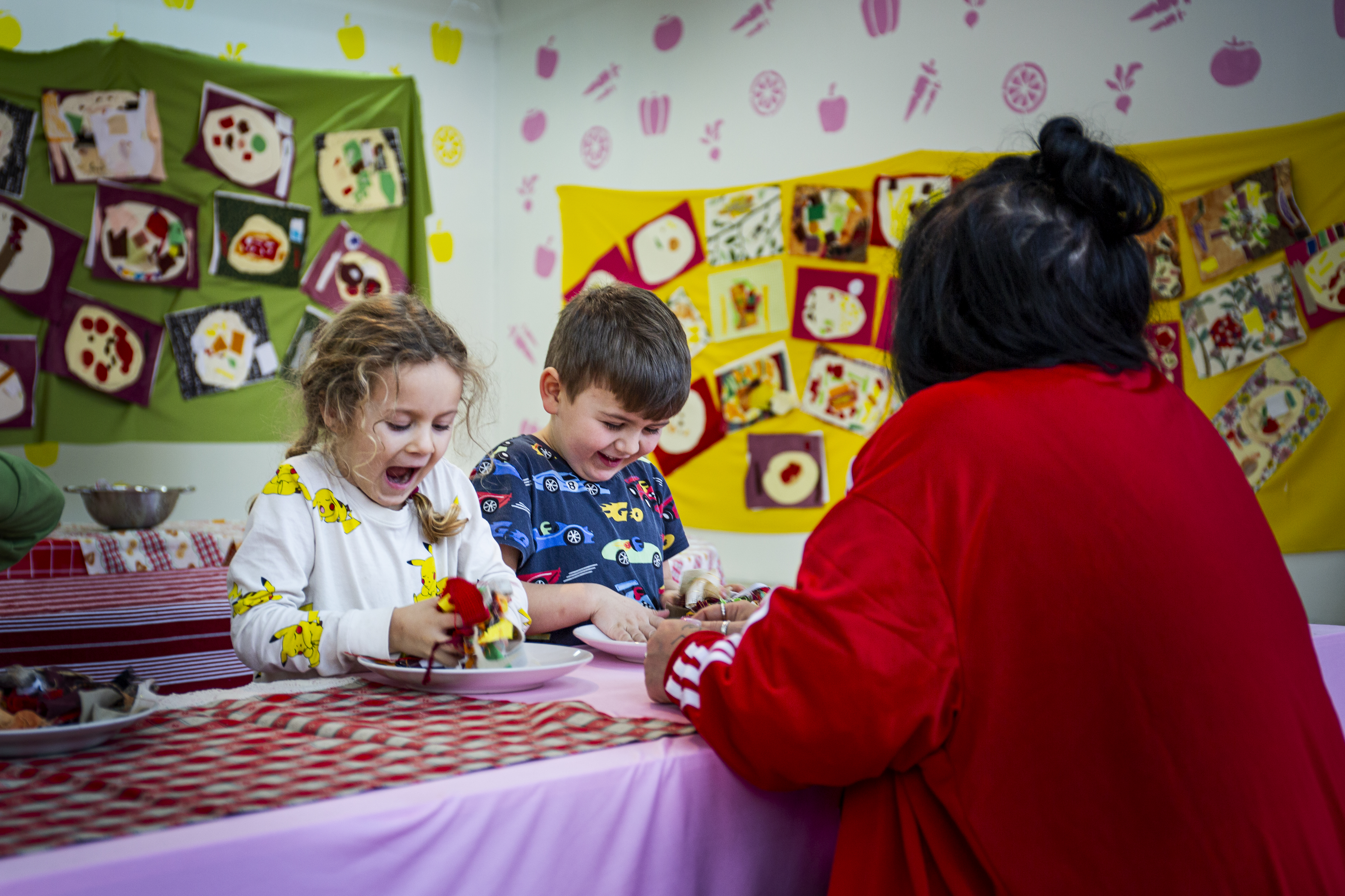 Två barn dekorerar tygmat vid ett bord under uppsikt av en vuxen. Rummet har konst på väggarna.