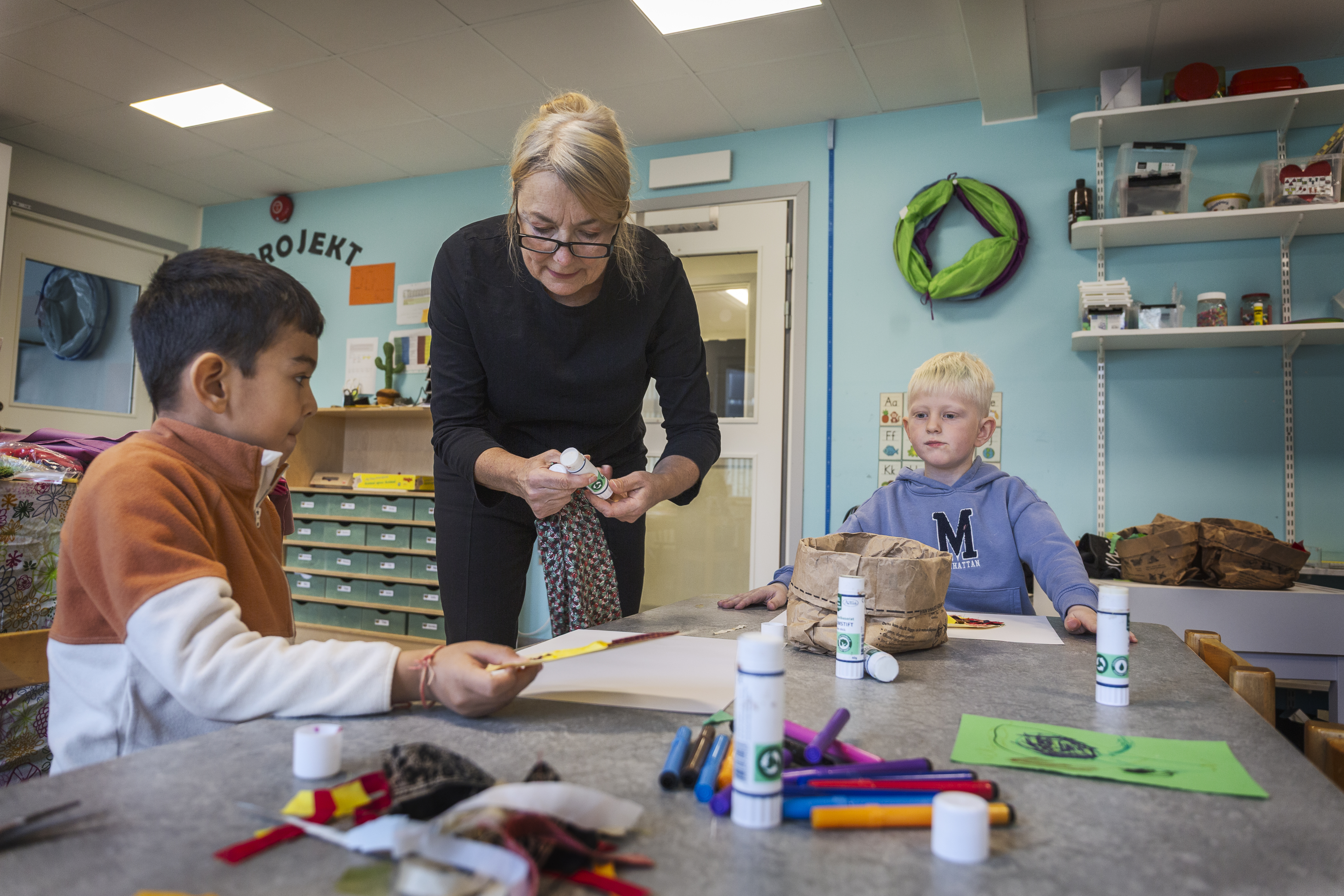 Birgitta Adolfsson hjälper två barn med sina konstprojekt vid ett klassrumsbord.