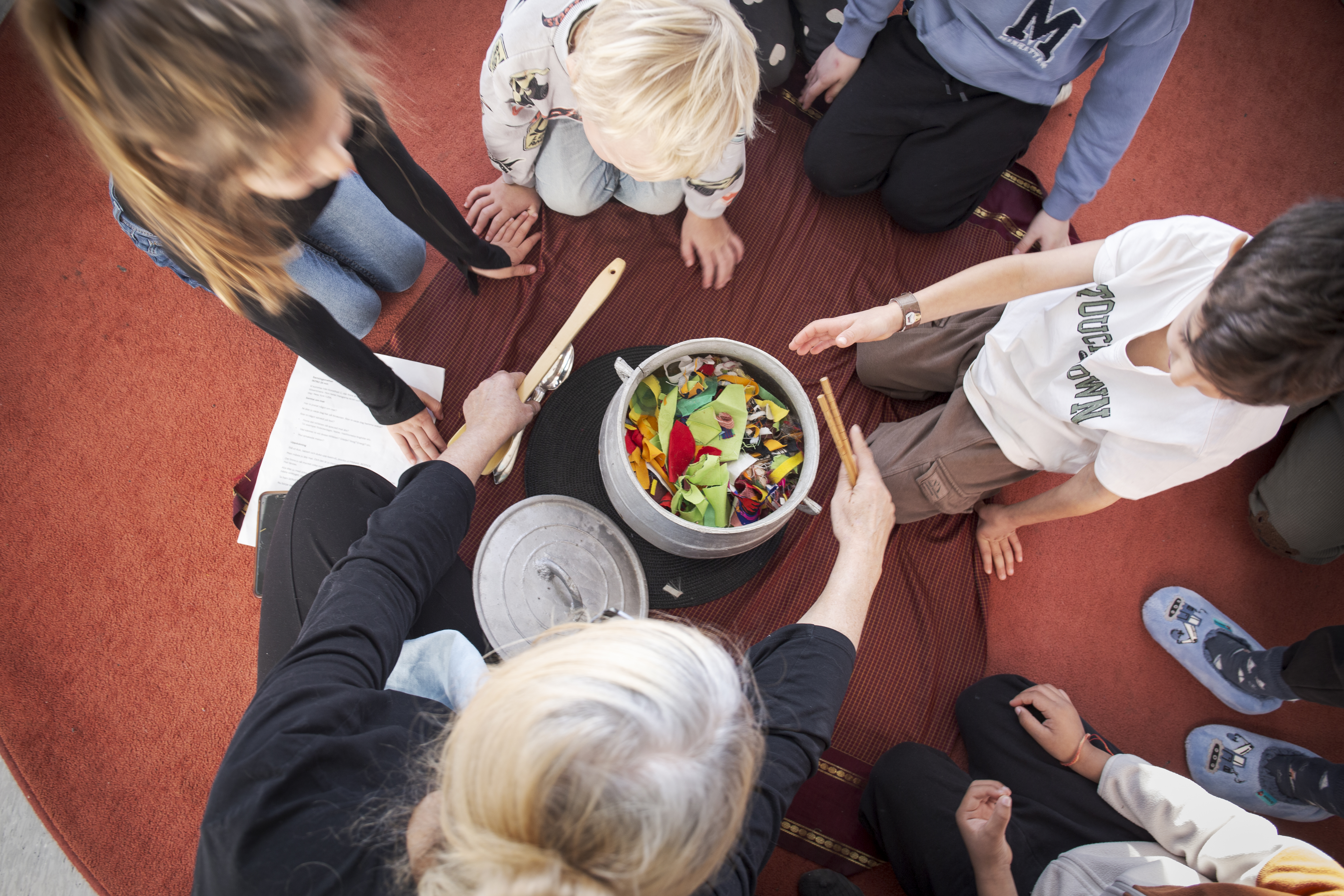Fyra barn och en vuxen sitter på en röd matta runt en gryta med låtsasmat.