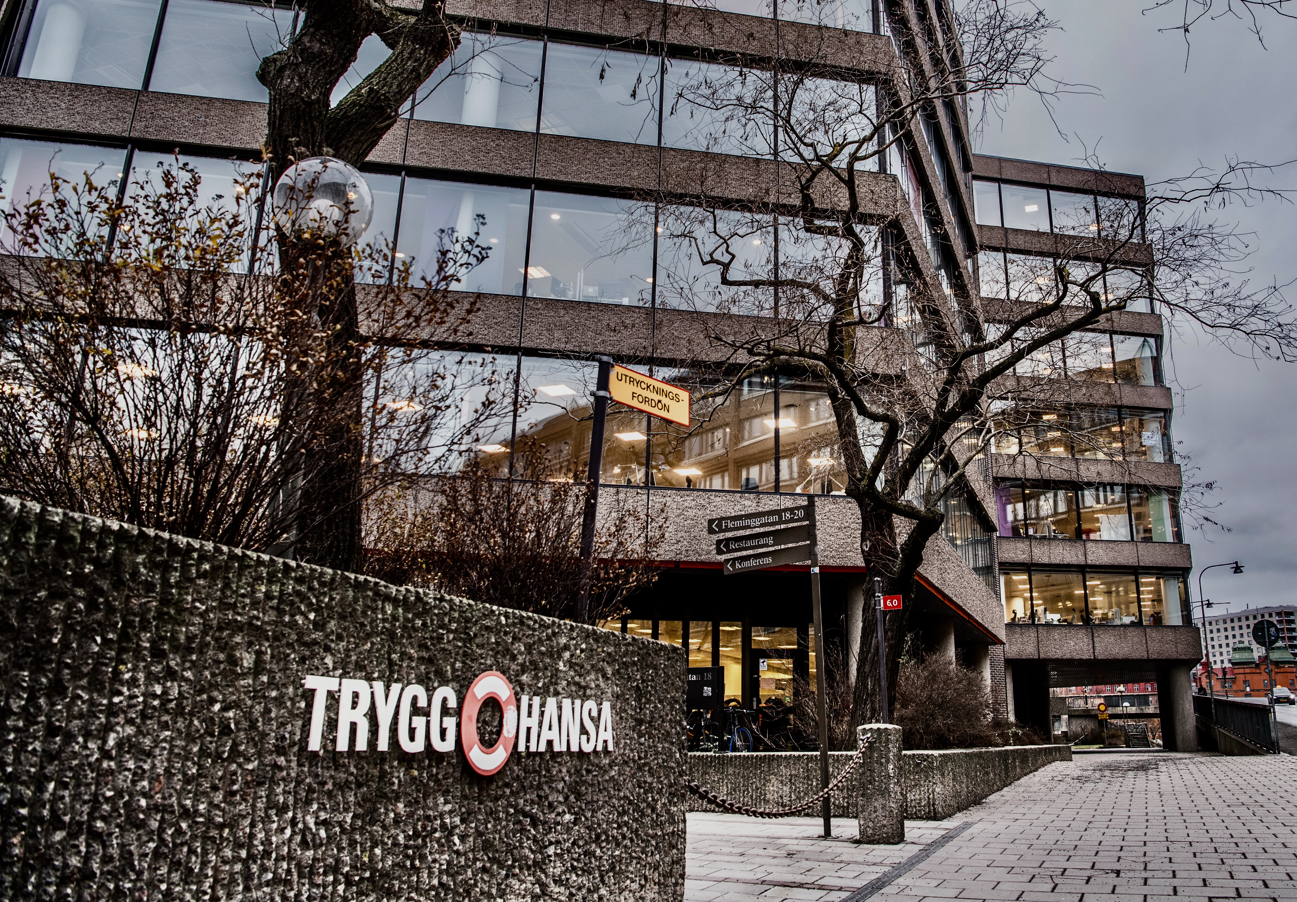 Trygg Hansas kontor med stora fönster, träd utan löv och en skylt med texten "TRYGG-HANSA" på en stenmur. Gatuskyltar och cyklar vid entrén.