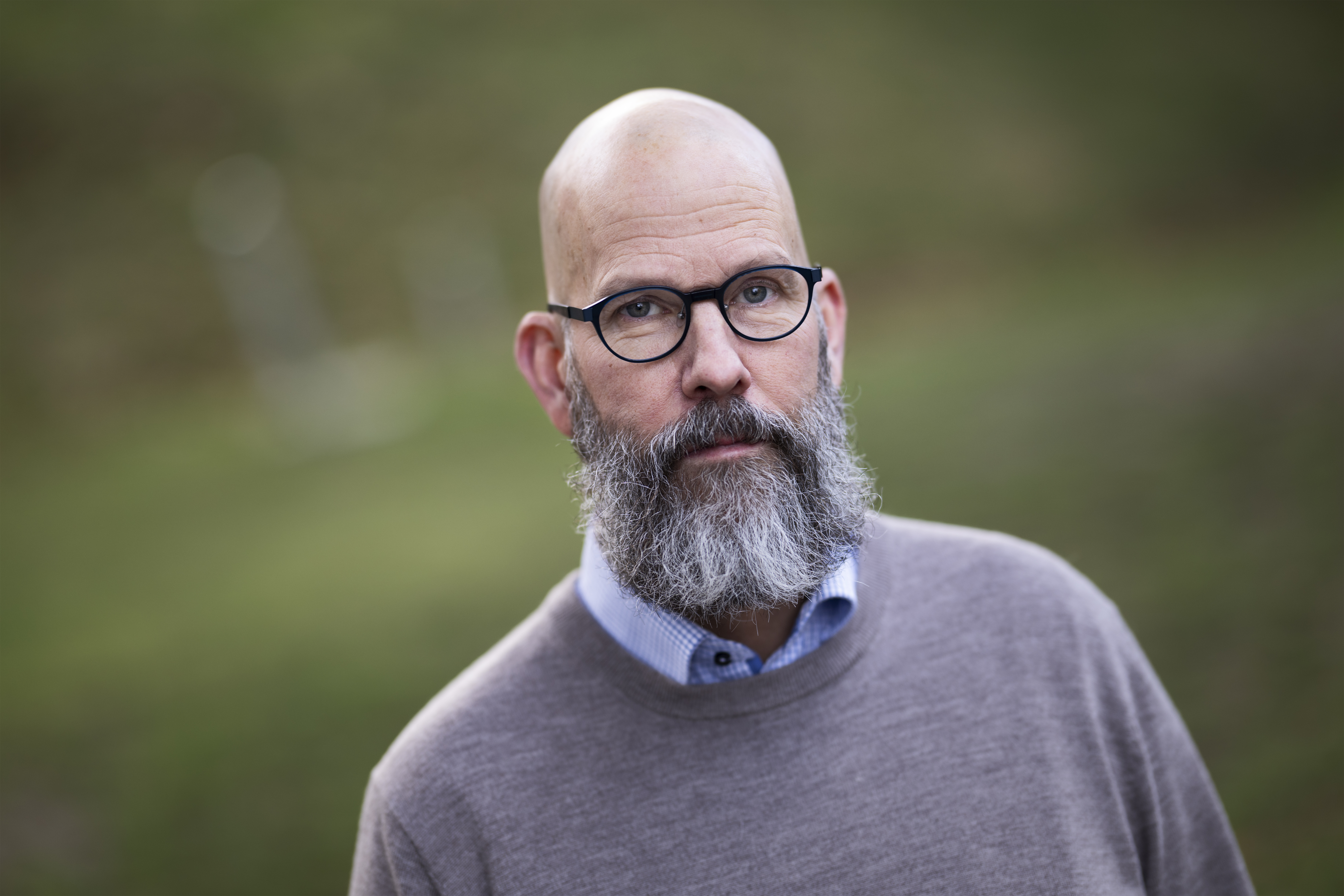 En man med grått skägg och glasögon, står utomhus framför en suddig grön bakgrund.