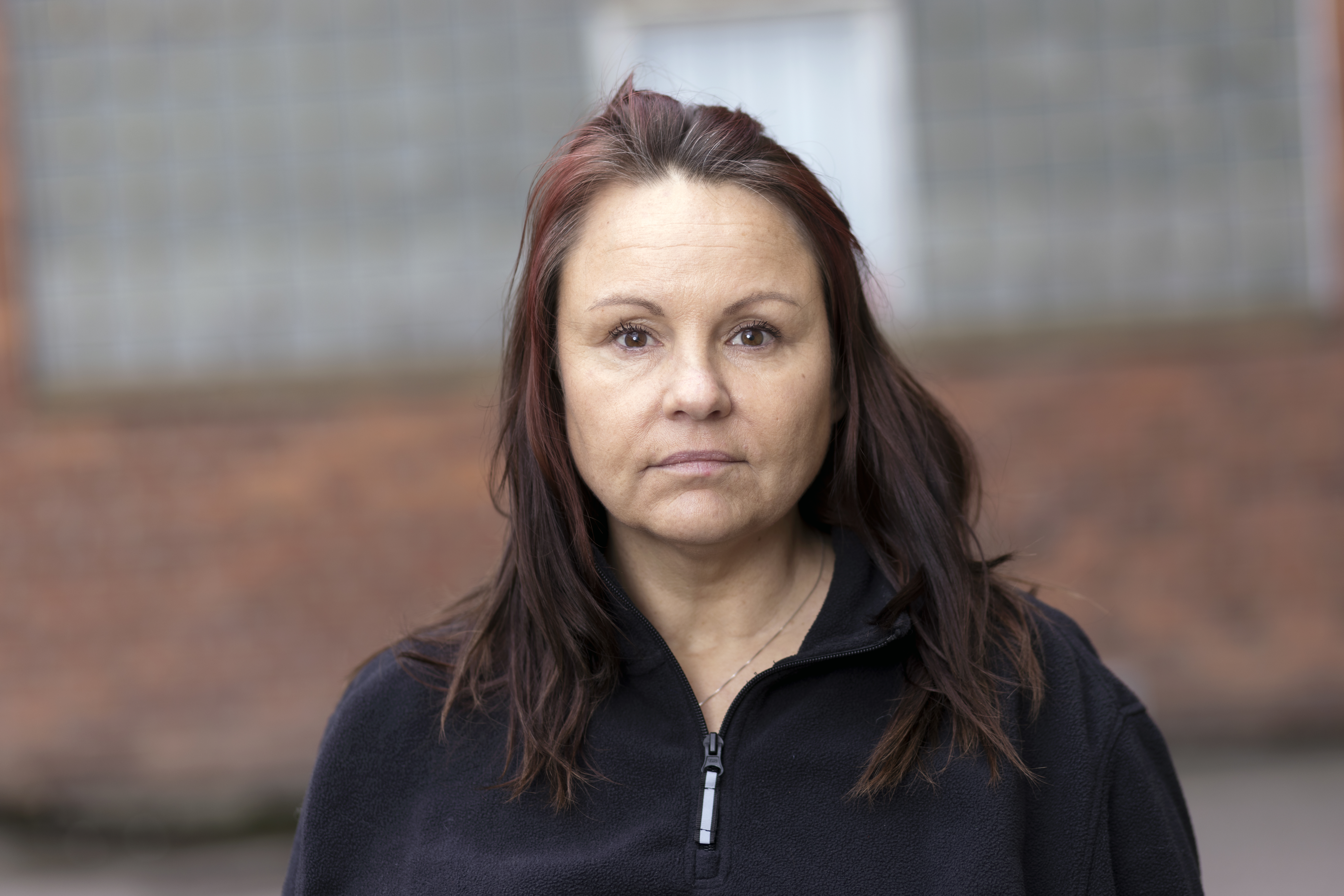 Jessica Axelson med långt brunt hår och svart topp med dragkedja står framför en tegelvägg utomhus och tittar in i kameran.