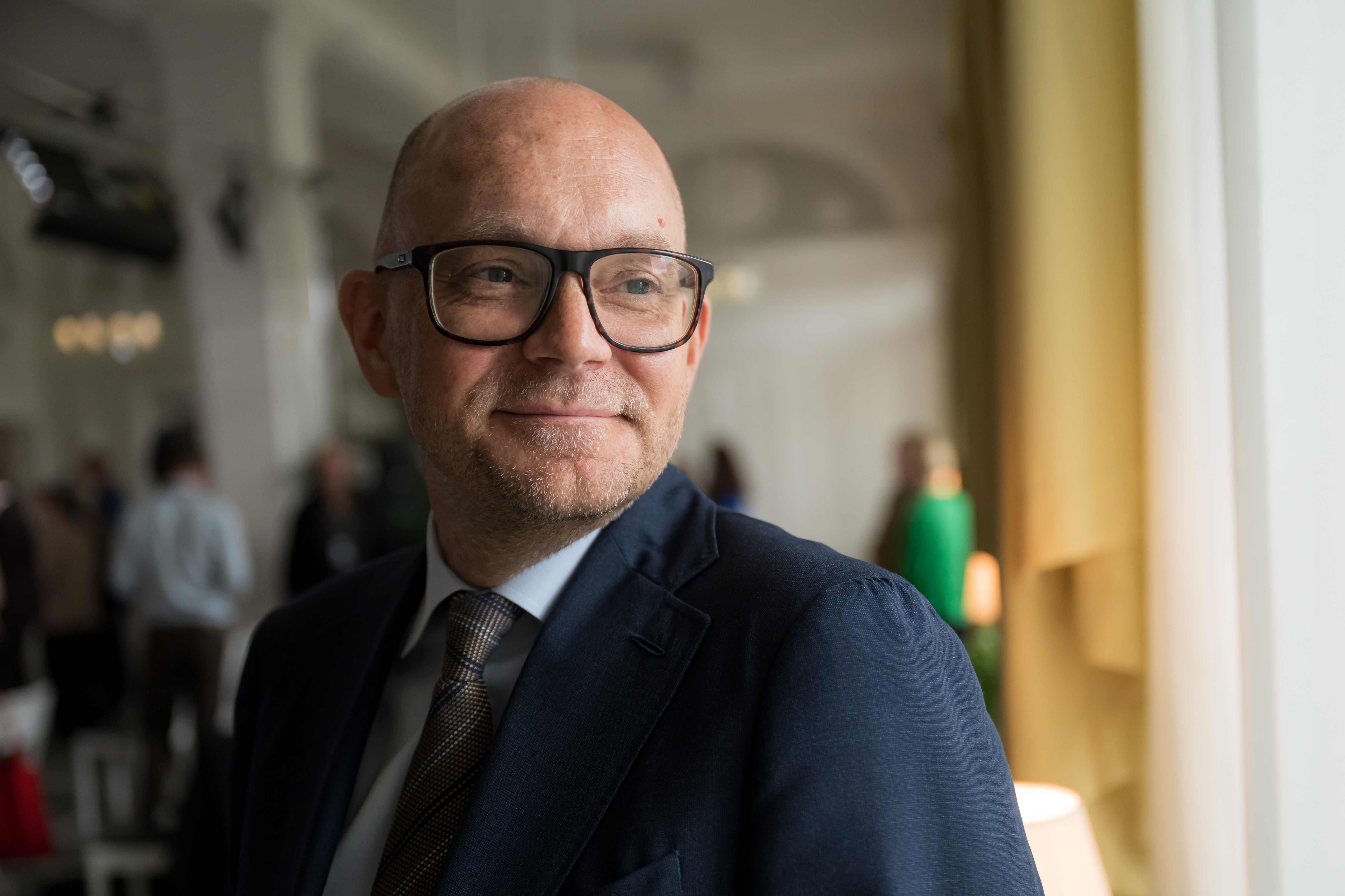 En man med glasögon och kostym står inomhus vid ett fönster och tittar åt sidan, flera personer syns i bakgrunden.