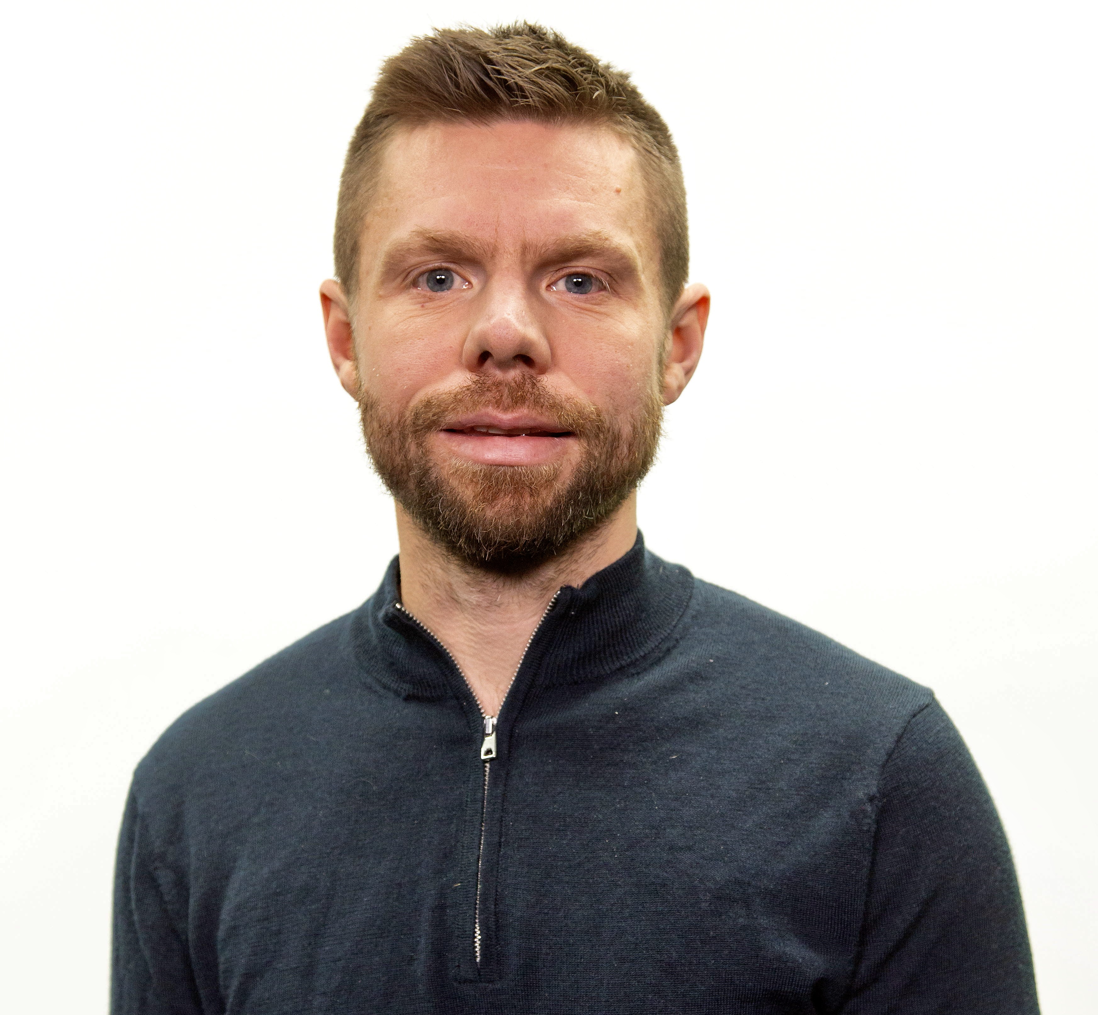 Henrik Örnlind med kort brunt hår och skägg har på sig en mörk tröja med dragkedja och står mot en vit bakgrund.