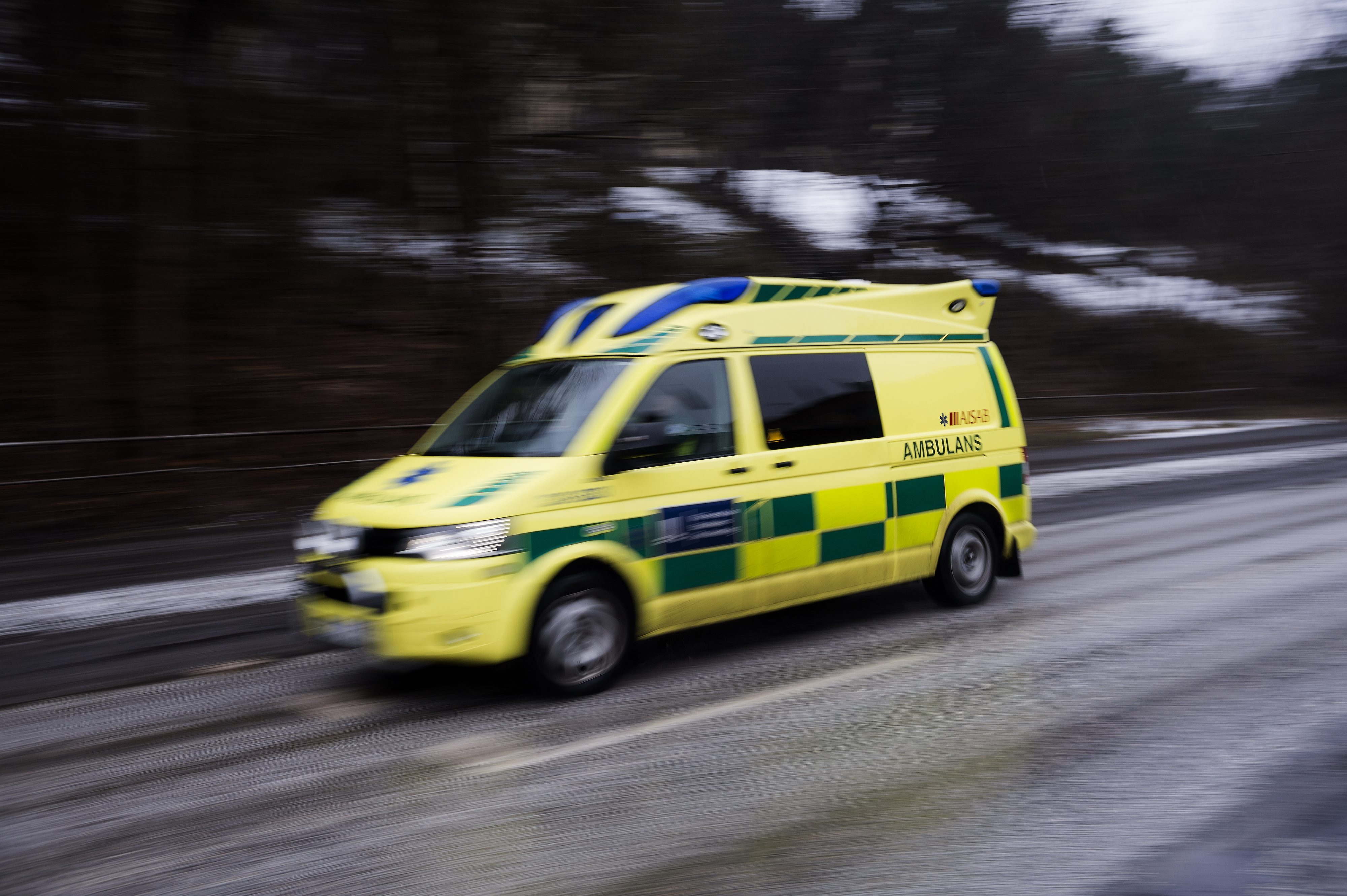 En gul och grön ambulans kör på en våt väg, träd och bakgrund är suddiga vilket visar att fordonet är i rörelse.