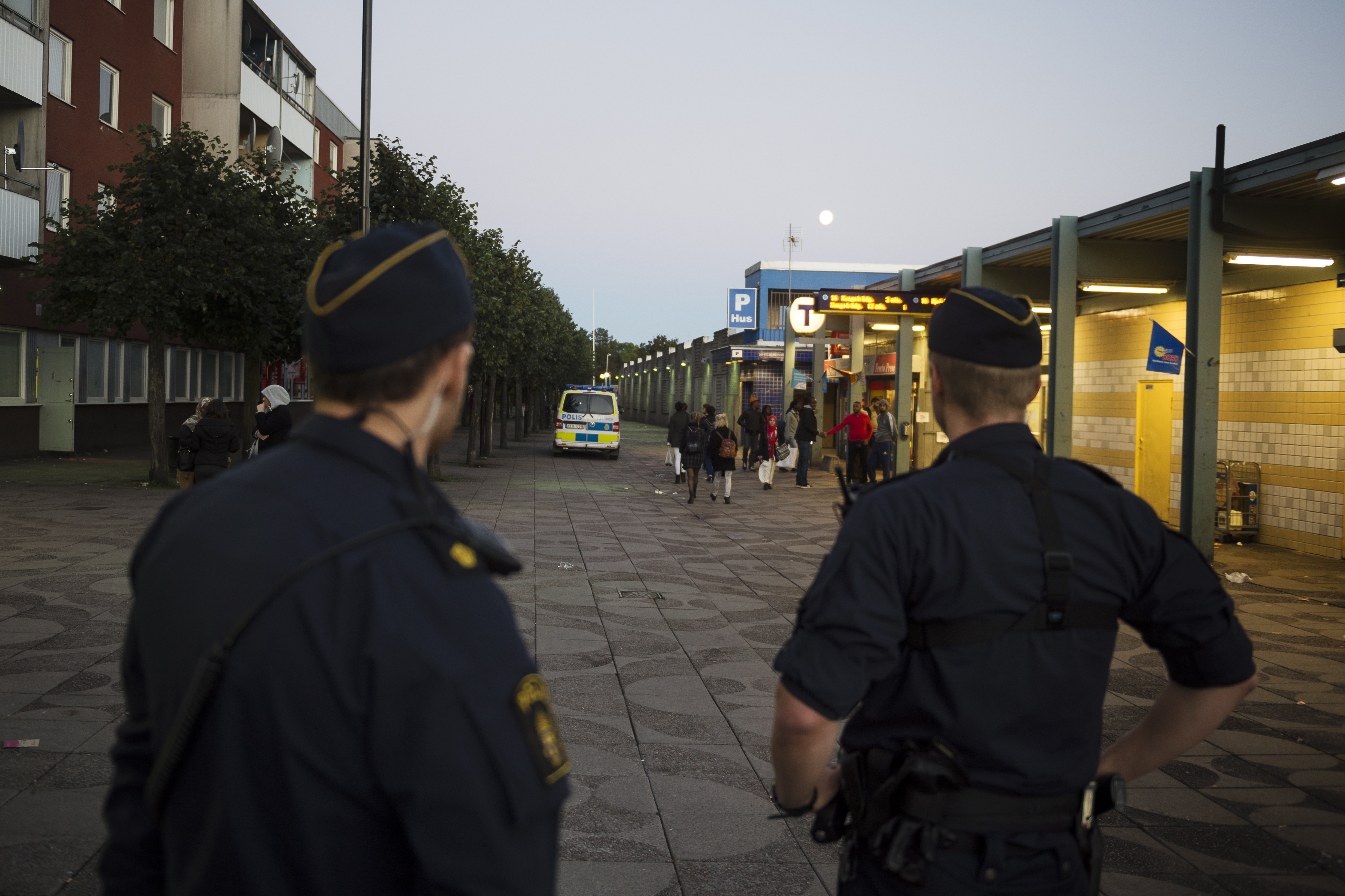 Två poliser övervakar ett torg i Rinkeby nära en lägenhetsbyggnad, med en polisbil och fotgängare i bakgrunden.