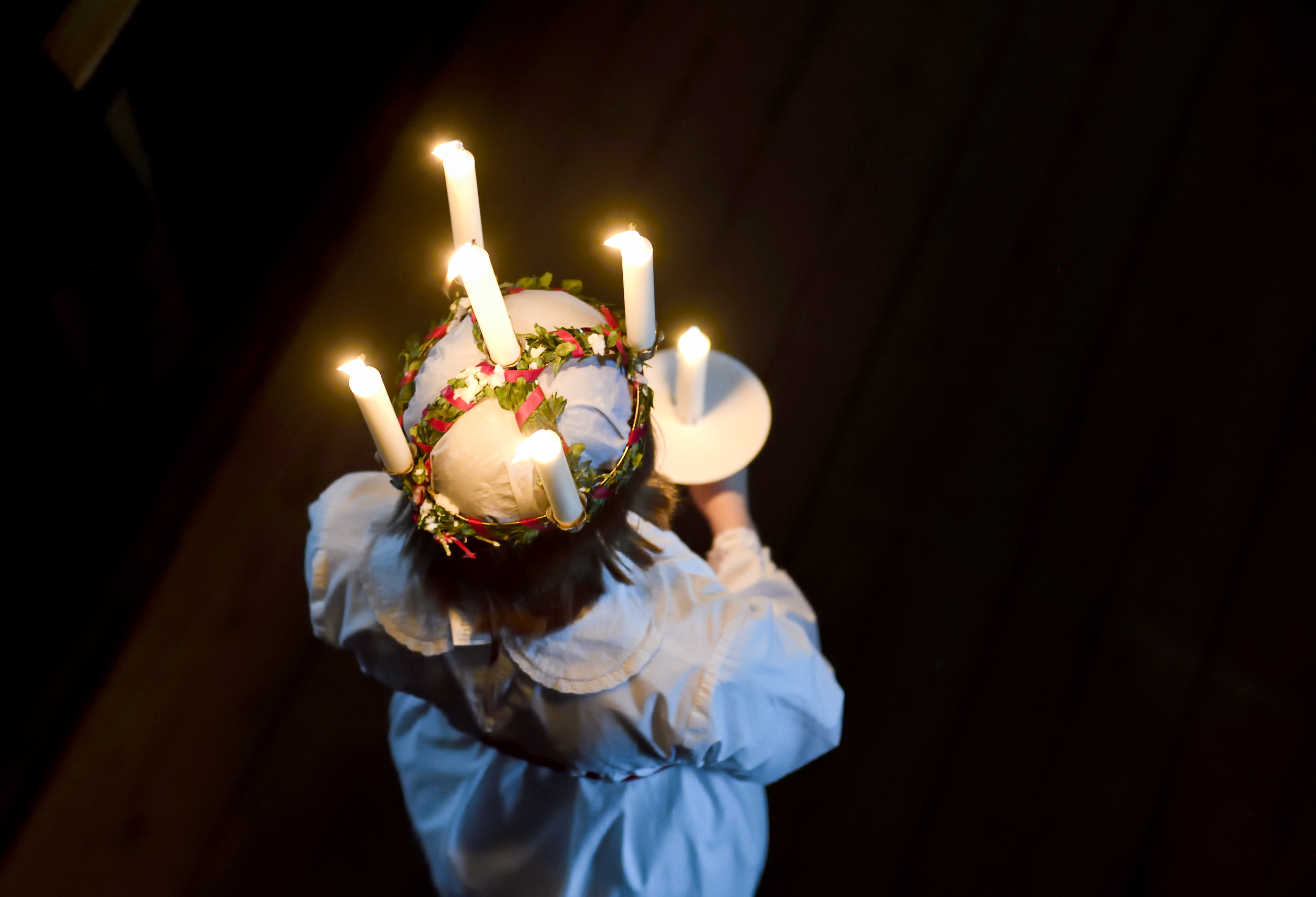 En lucia har på sig en vit dräkt och en krona med tända ljus och blommor, håller ett vitt ljus och står på ett trägolv.