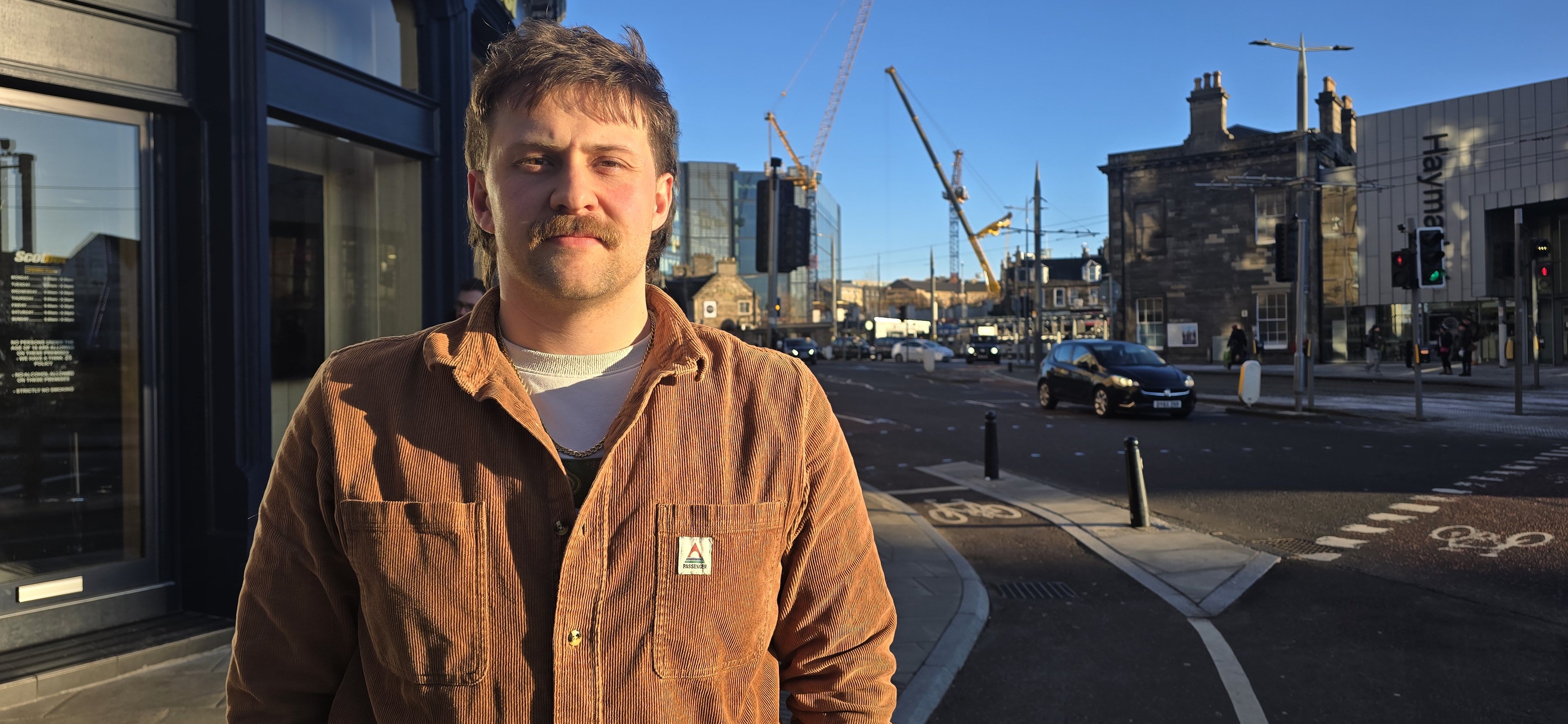 En man med mustasch står på ett gatuhörn i en stad. I bakgrunden syns byggnader, byggkranar och en bil som passerar.